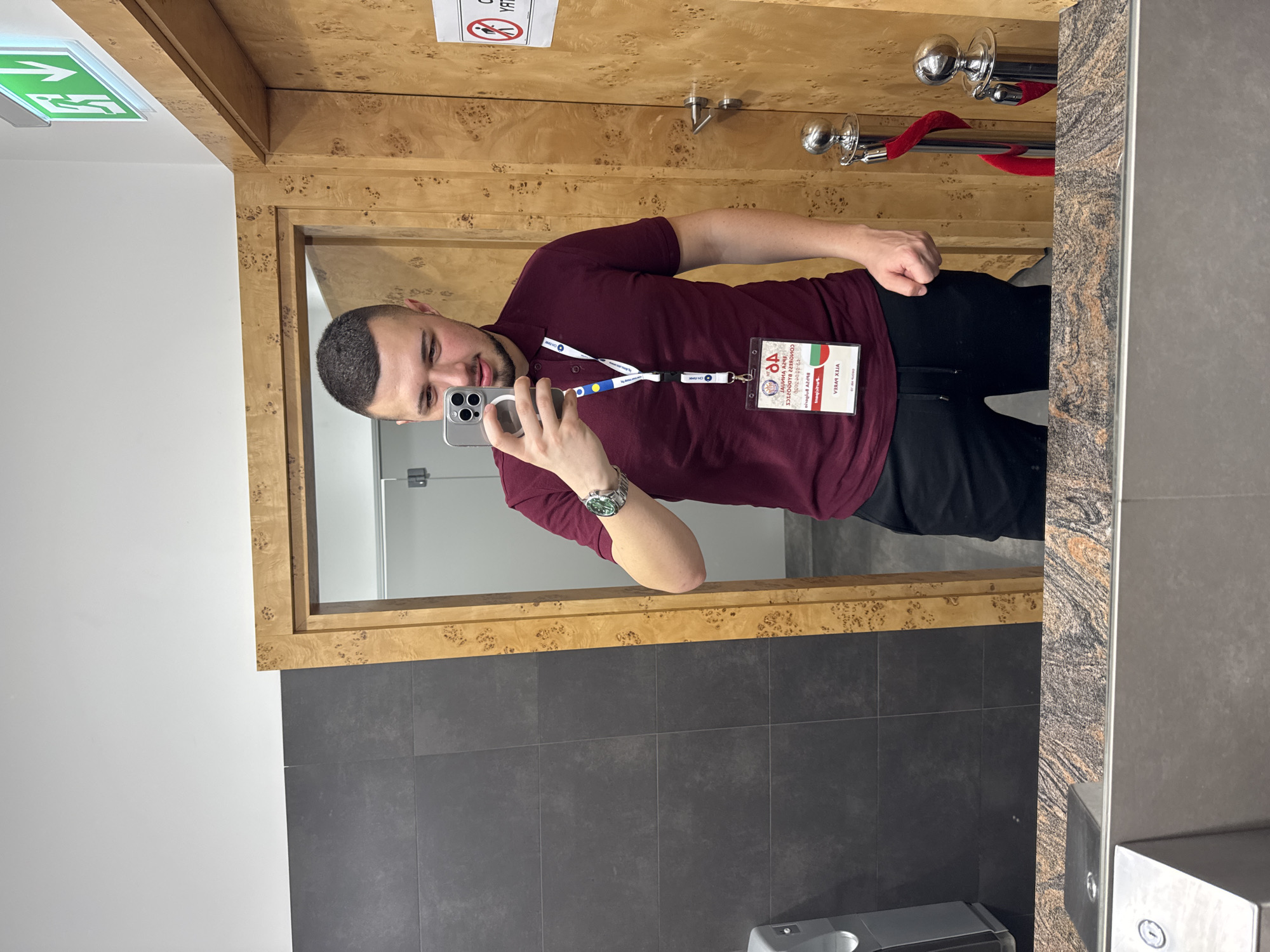 A man is taking a selfie in a bathroom. He is wearing a maroon polo shirt, black pants, and a watch on his left wrist. He has a lanyard around his neck with an identification badge. The bathroom has a dark gray tiled wall and a light wood-framed mirror. There is a red velvet rope behind him and a green emergency exit sign visible in the upper left corner. The reflection in the mirror shows a white tiled bathroom stall. Based on the identification badge with the letters "OA", it can be assumed this picture was taken in Opera Nova, Bydgoszcz, Poland.