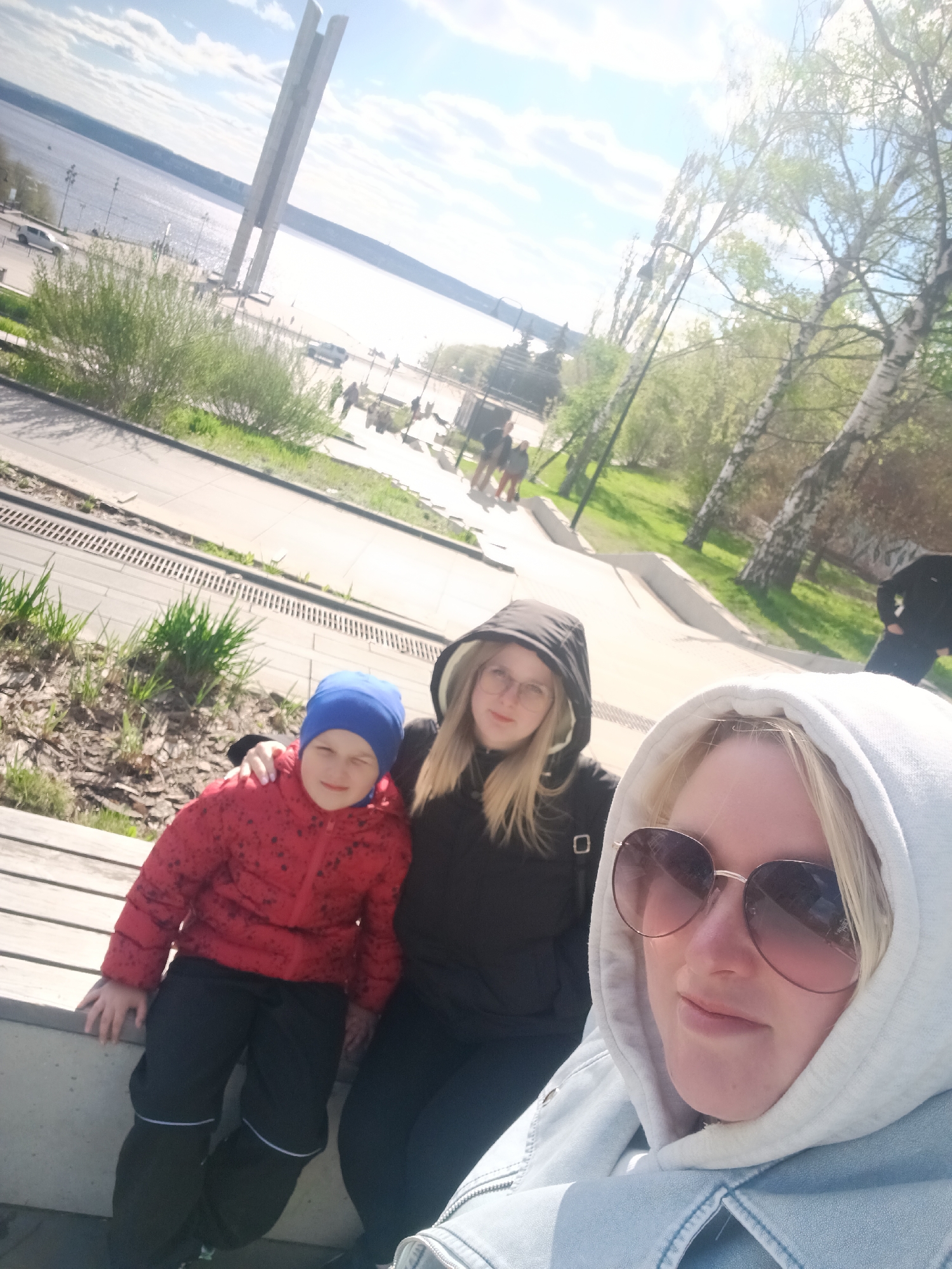 Three people are in the image. A child in a red jacket and blue hat sits to the left, next to a person in a black hooded jacket and glasses. The person taking the photo, wearing sunglasses and a light-colored hooded jacket, is positioned on the right. The background shows a city landscape. There's a body of water with a monument visible in the distance. The location is Izhevsk, Russia. A pathway with greenery and trees is to the right.