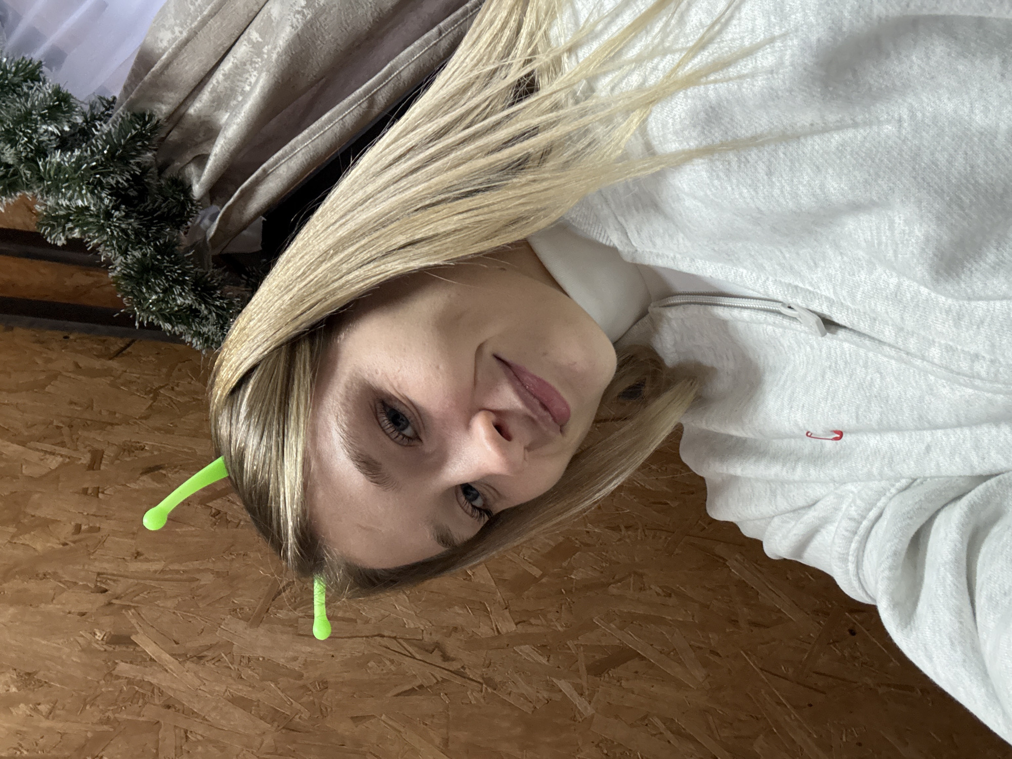 A woman with blonde hair, wearing a light grey hoodie and green antennae on her head, looks at the camera. The background appears to be a wooden surface. A Christmas garland hangs in the top-left corner. The image was taken in Прикамский, Russia.