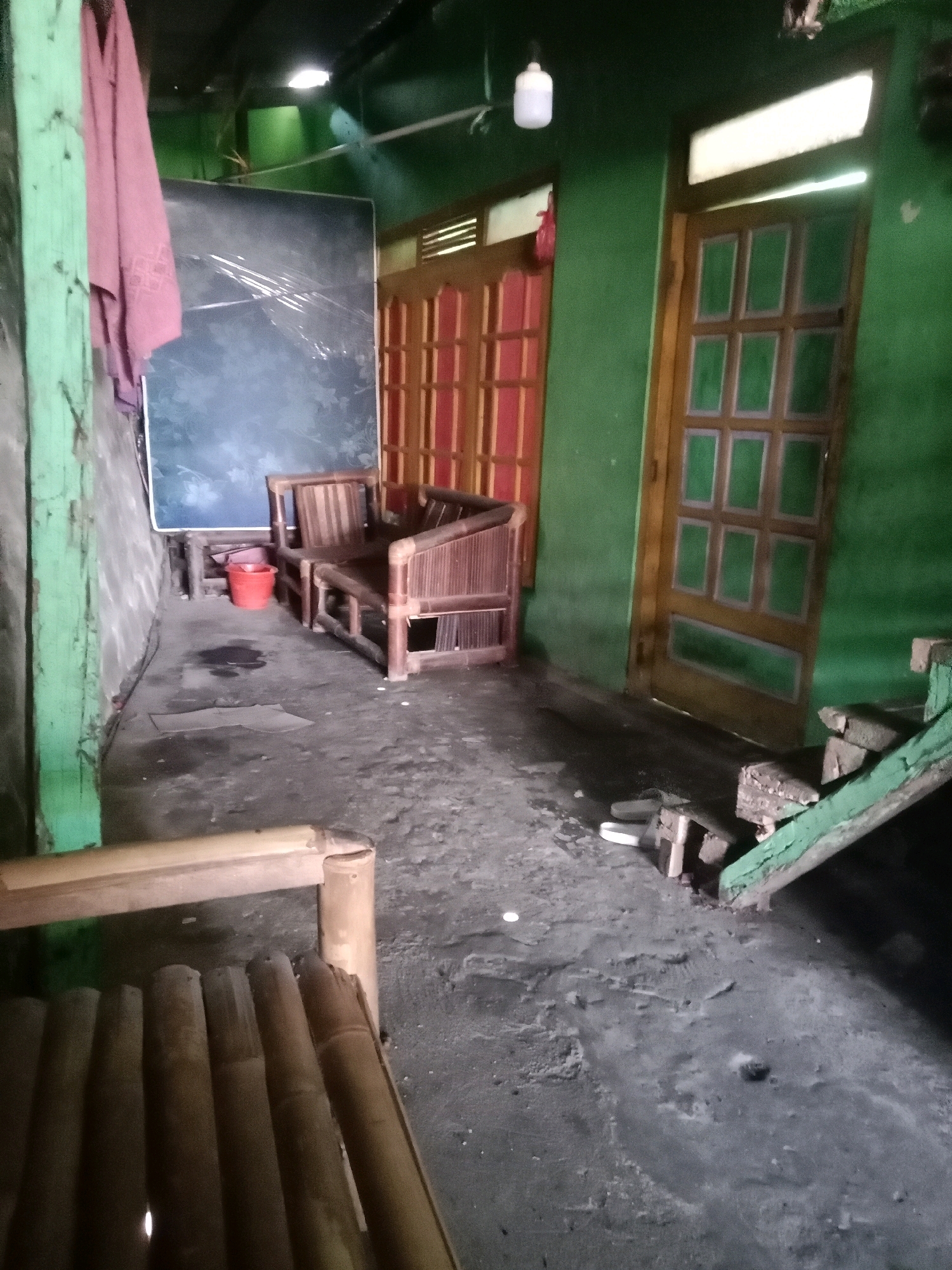 The image depicts a dimly lit, cluttered, outdoor or semi-outdoor space, possibly a back porch or alleyway, located in Adipuro, Indonesia. The walls are painted a vibrant, uneven green. In the foreground, a portion of a bamboo bench or chair is visible, made of rough-hewn bamboo poles. The floor is a rough, textured surface, appearing to be concrete or packed earth, with several dark, wet-looking stains and some scattered debris, including a piece of cardboard.
To the left, a green-painted vertical wooden post is prominent, with a pink towel or piece of fabric hanging from it. Behind this, a large, dark, rectangular object, possibly a mattress or board, leans against the wall. A small, bright red bucket sits on the floor near the object.
Further into the scene, two bamboo chairs, likely a set, are positioned centrally. They are made of woven bamboo strips and thicker bamboo poles. Beyond the chairs, there are two wooden doors with glass panes. The door on the left is painted red within the panes and has a wooden frame. The door on the right is framed in wood and has panes with green-colored glass or material. A narrow, angled wooden structure painted green, possibly part of a staircase or railing, is seen on the right side of the image, partially obscuring the door.
Above, the ceiling appears to be dark, with a white ceiling fan and a simple white light fixture visible. The overall impression is of a utilitarian, somewhat neglected space.