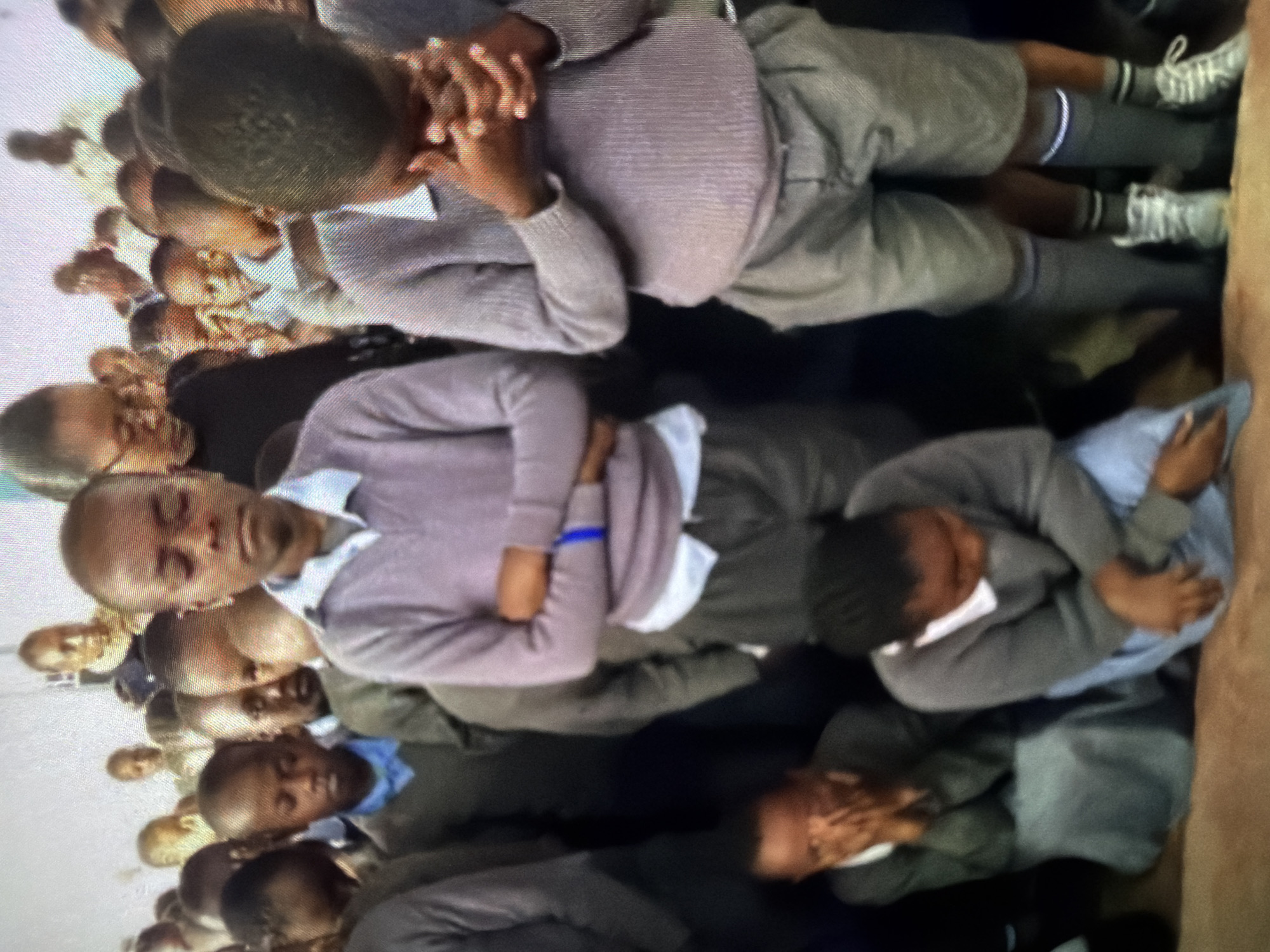 A group of children, likely students, are gathered together indoors, possibly in a classroom or assembly hall. Many of them are wearing similar school uniforms, consisting of light-colored sweaters over white collared shirts and grey pants or skirts. Some of the children have their hands clasped in front of their faces, appearing to be praying or in deep thought. The children are of various ages, and the scene suggests a solemn or reflective moment. The background is out of focus, but appears to be a plain wall, indicating an indoor setting.