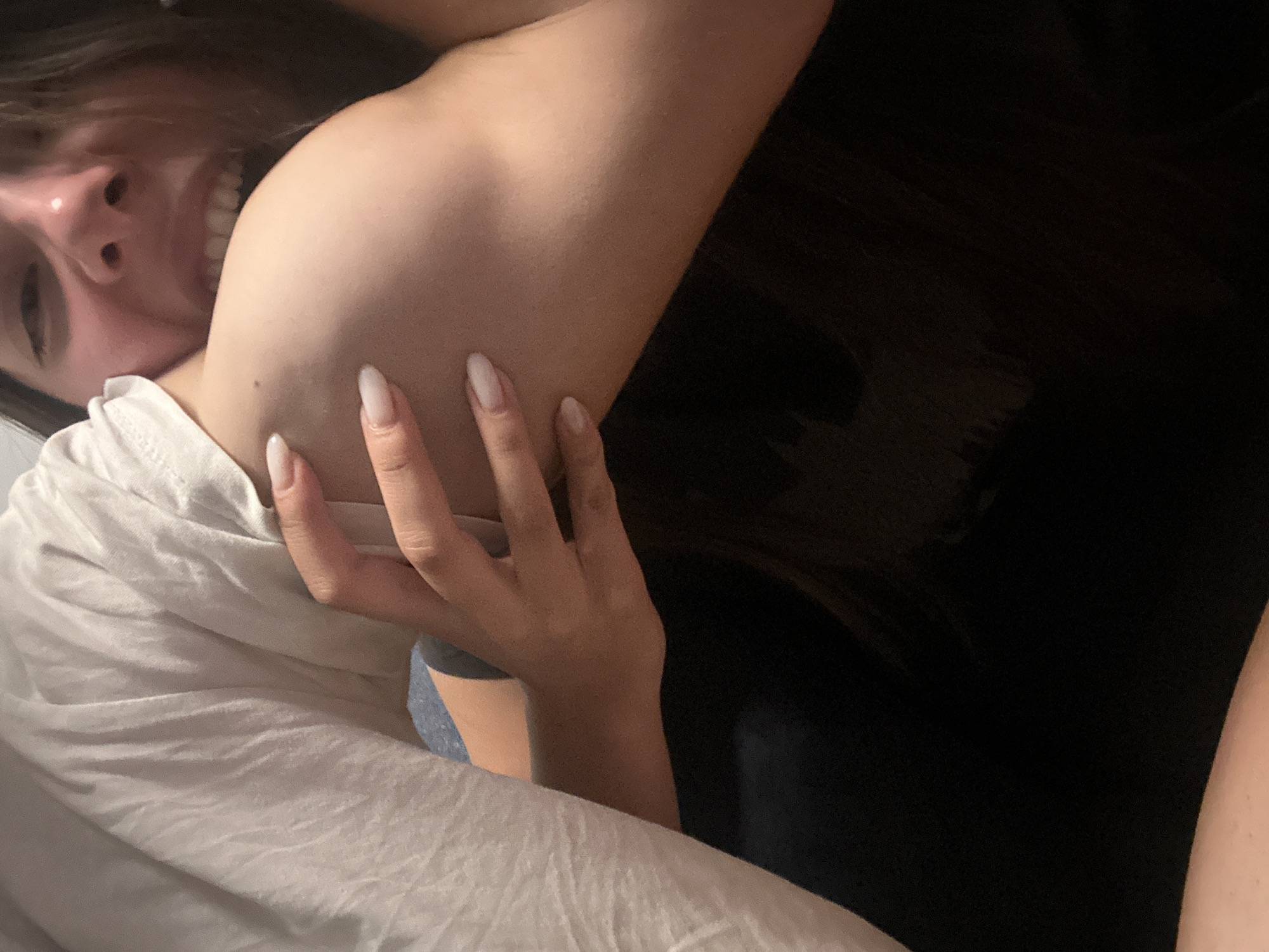 The image is a close-up, slightly blurry shot focusing on a person's shoulder and face. The person on the right is biting the shoulder of the person on the left, who is wearing a white t-shirt. The person biting has their mouth open, revealing teeth, and their eyes are looking towards the camera. The hand of the person being bitten is visible, resting on the shoulder. The background is dark and out of focus. The image appears to be taken indoors, possibly in a residential setting. There are no visible elements that indicate a specific location in Sofia, Bulgaria.