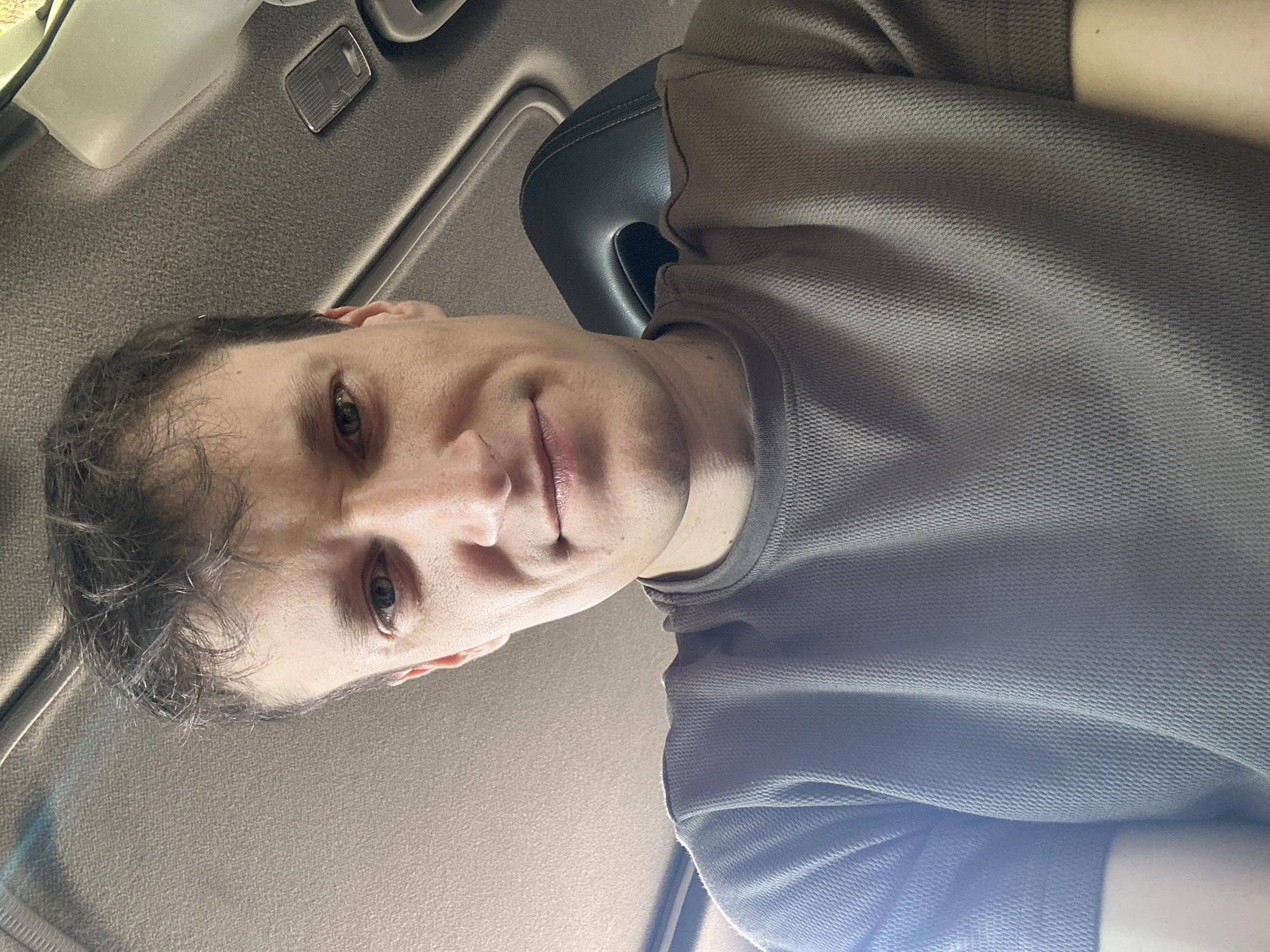 A low-angle, selfie-style shot shows a man looking up towards the camera from inside a vehicle. The man has short, messy brown hair and is wearing a brown, textured t-shirt. His face is partially obscured by shadows, but his eyes are visible, looking directly at the camera. The interior of the car is visible above him, with the textured headliner and part of a light fixture in the ceiling. A black headrest is visible to the right of the man's head. The lighting suggests it is daytime, with natural light filtering in from outside. The background does not provide any information about the specific location within Makiv, Ukraine.