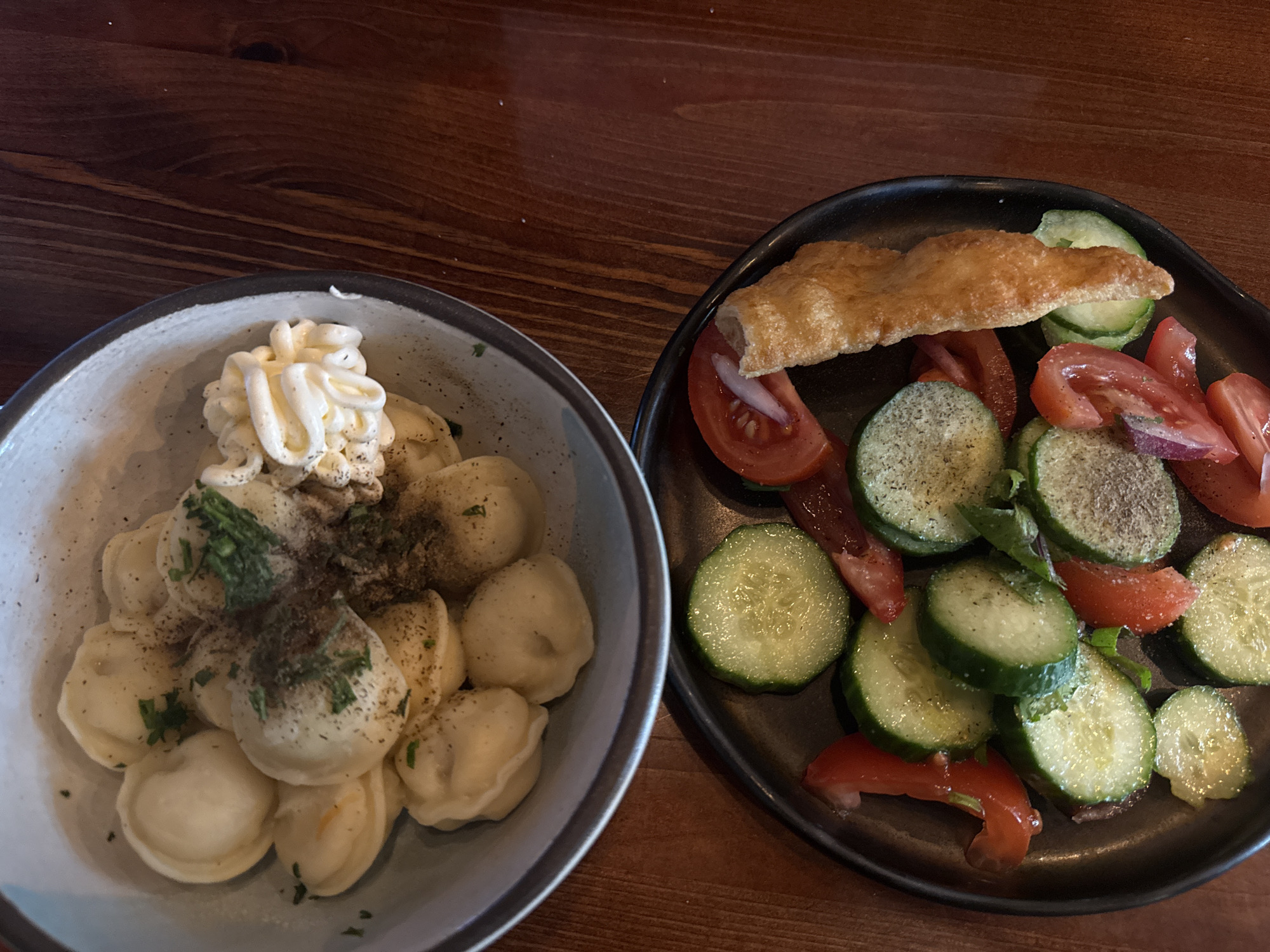 The image shows a meal served on a wooden table. On the left, there's a bowl of pelmeni (dumplings) topped with a swirl of mayonnaise and sprinkled with herbs and pepper. To the right, there is a plate with sliced cucumbers, tomatoes, and what appears to be a piece of bread or pastry. While the image itself does not specify the location as Moscow, Russia, the dish is commonly consumed within its borders.
