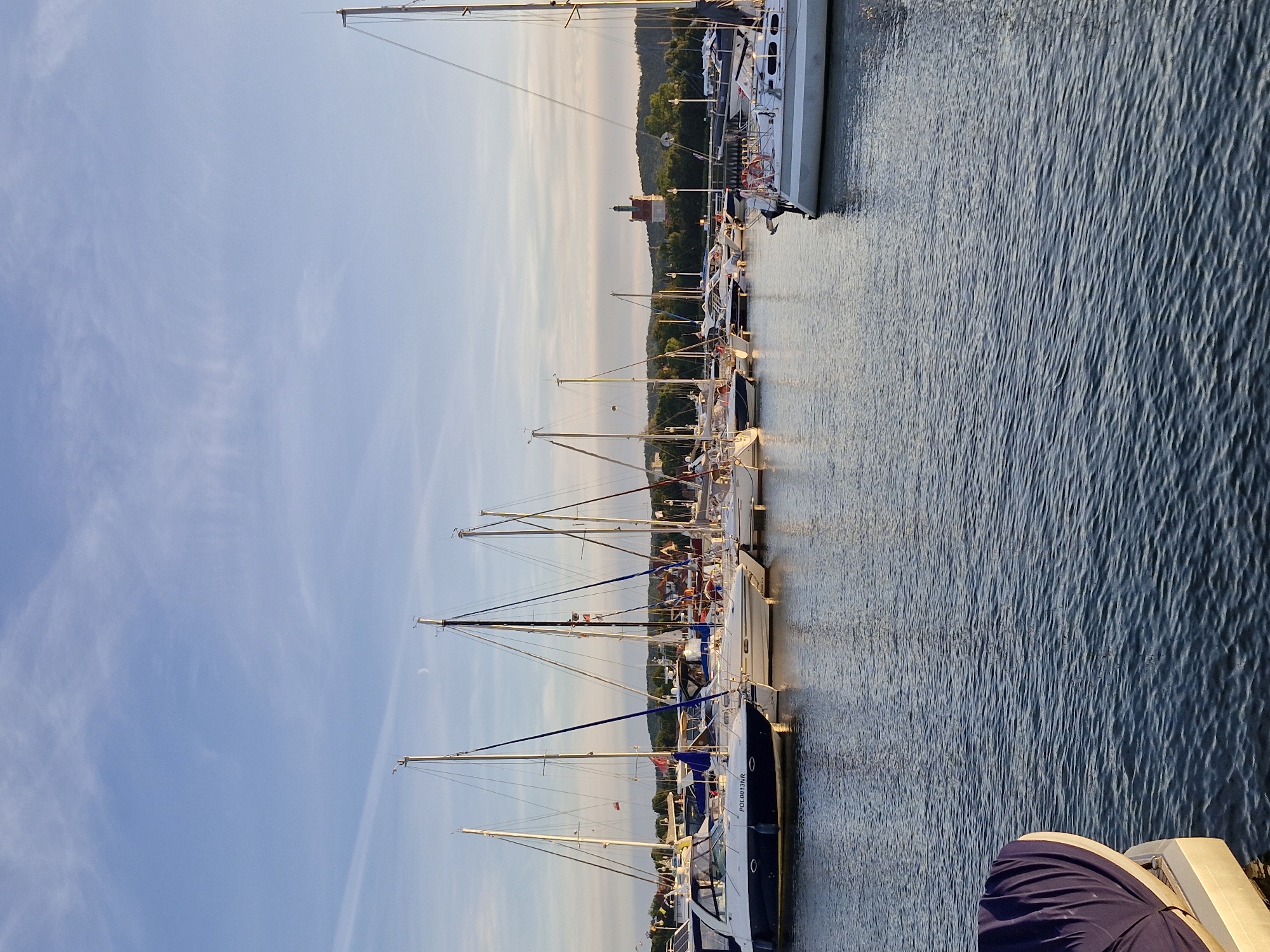 The image shows a marina in Sopot, Poland, with several sailboats docked. The sky is a clear blue with some wispy clouds. The sun is low in the sky, casting a warm glow on the scene. In the background, there are trees and buildings, including a distinctive tower that appears to be a lighthouse or observation tower. The water in the marina is calm and reflects the sky and the boats. One boat in the foreground has the registration "POL0013NR" visible.