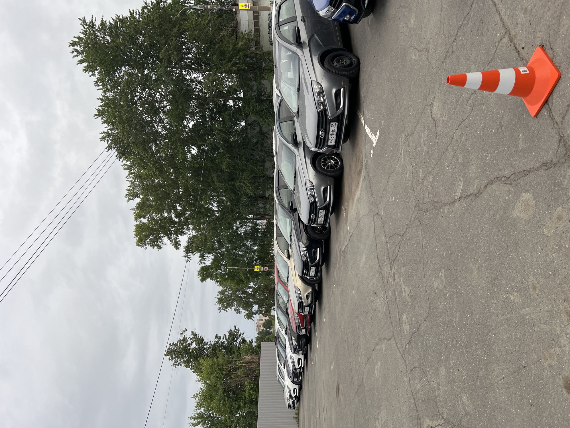 The image presents an outdoor asphalt parking lot located in Izhevsk, Udmurt Republic, Russia, under an overcast sky. A linear arrangement of twelve passenger vehicles, predominantly Lada Vesta and Granta models, are parked diagonally, facing the viewer. The cars exhibit a range of colors including white, black, various shades of grey, silver, red, and gold. Russian license plates with the regional code '18' (Udmurt Republic) are discernible on multiple vehicles, such as E431MC 18, M532OC 18, T197HT 18, and X024BA 18. The asphalt surface of the lot is cracked and shows faded white ground markings. A grey corrugated metal fence runs along the left side of the parking area, while a dense line of green deciduous trees forms the background. Faint urban buildings are visible above the treeline in the distance. A utility pole on the right supports a street lamp and Russian road signs, specifically a yellow triangular "Children" warning sign and a circular "40 km/h" speed limit sign. Multiple power lines traverse the upper portion of the sky. A single orange and white striped traffic cone with a square base and a white identification sticker is positioned in the lower right foreground. No individuals are present in the scene.