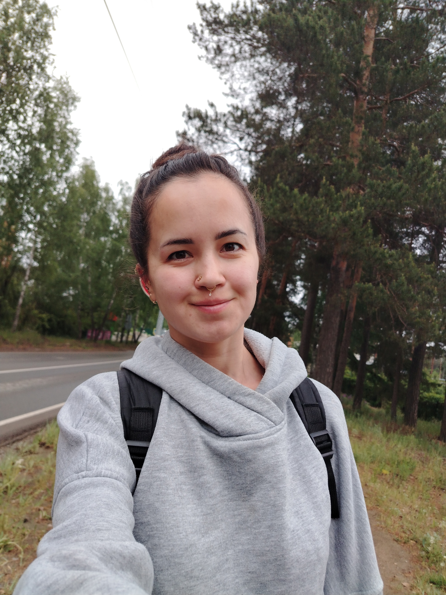 The image is a selfie of a woman with fair skin and brown hair pulled back into a bun. She has a nose ring and multiple earrings. She is wearing a light gray hoodie and a black backpack. She stands on the side of a paved road with green foliage on both sides. Tall pine trees are visible in the background. The location is likely Noviy, Russia, based on the image’s origin.