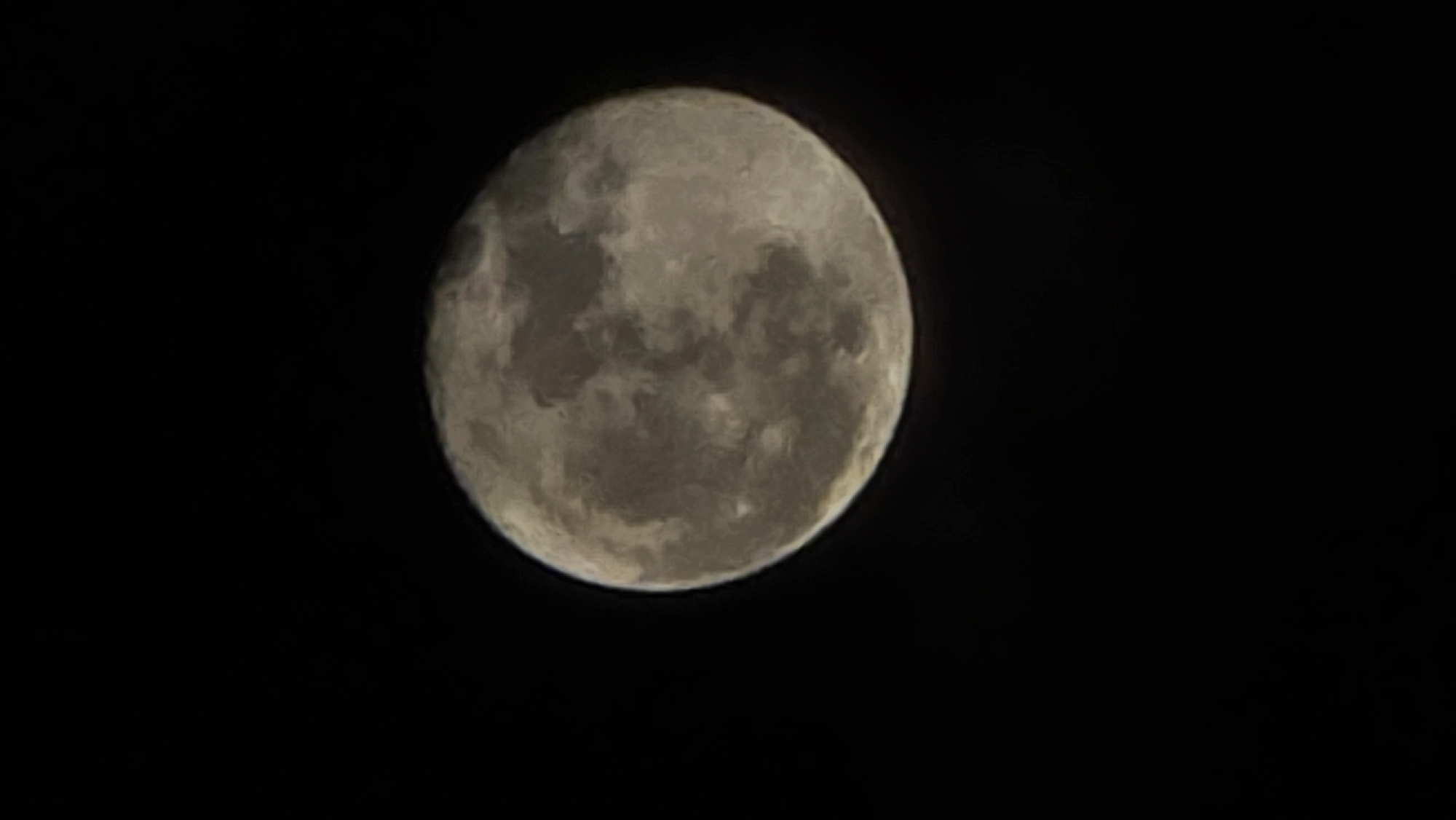 A close-up shot of the full moon in a black sky. The moon is round and illuminated, showcasing its surface texture with craters and shades of grey. The location is not discernible from this image.