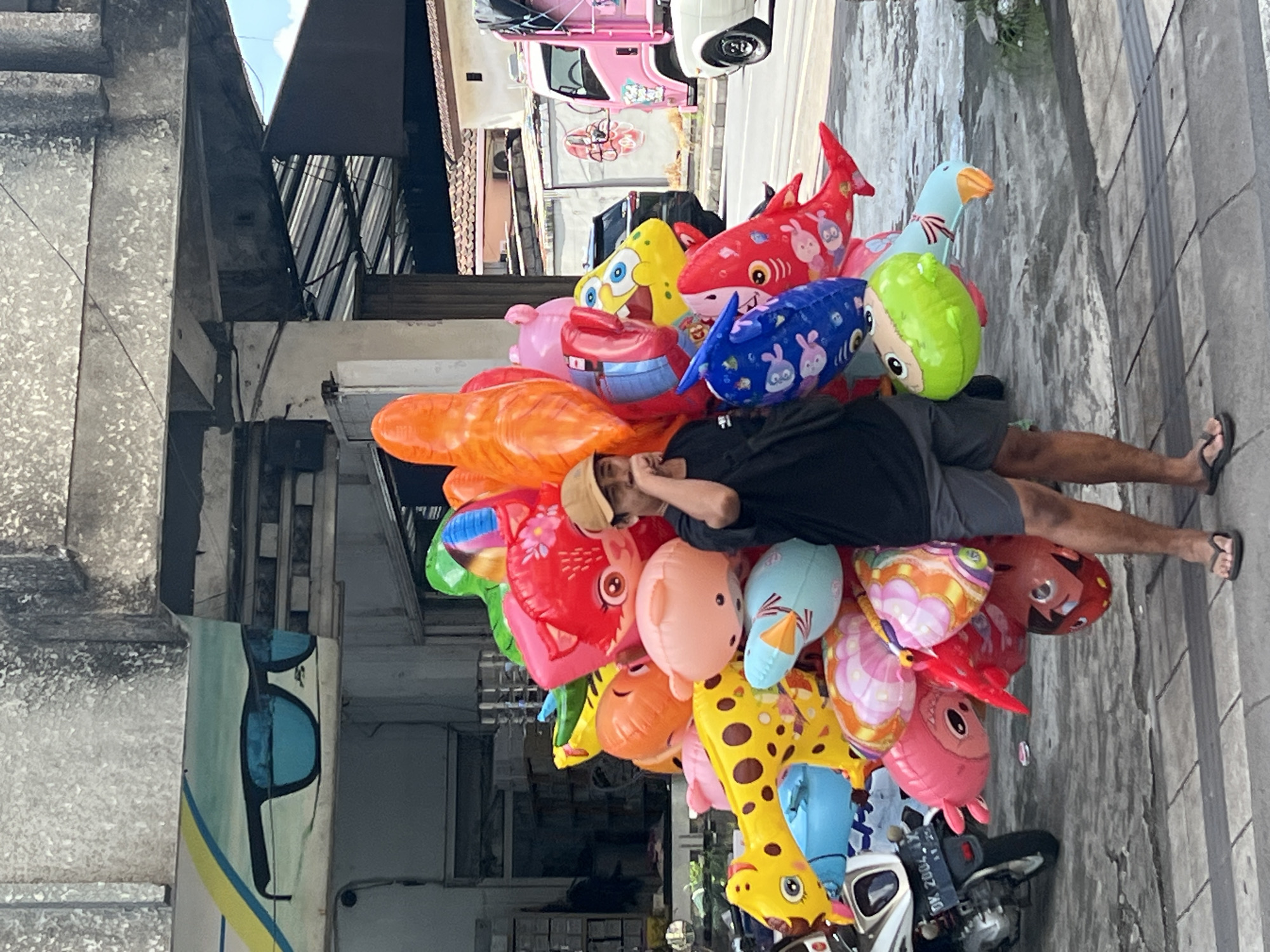 A man in Denpasar, Indonesia lies on a sidewalk with a large bouquet of colorful inflatable balloons. He is wearing a black t-shirt, gray shorts, and a tan hat. He is lying on his side, using his hand as a pillow. The balloons are various shapes and colors, including animals like sharks, a goose, and a giraffe, along with other cartoon characters. A scooter is parked nearby. Behind him is a building with a mural of sunglasses. Cars and other buildings are visible in the background. The sidewalk is made of gray tiles.