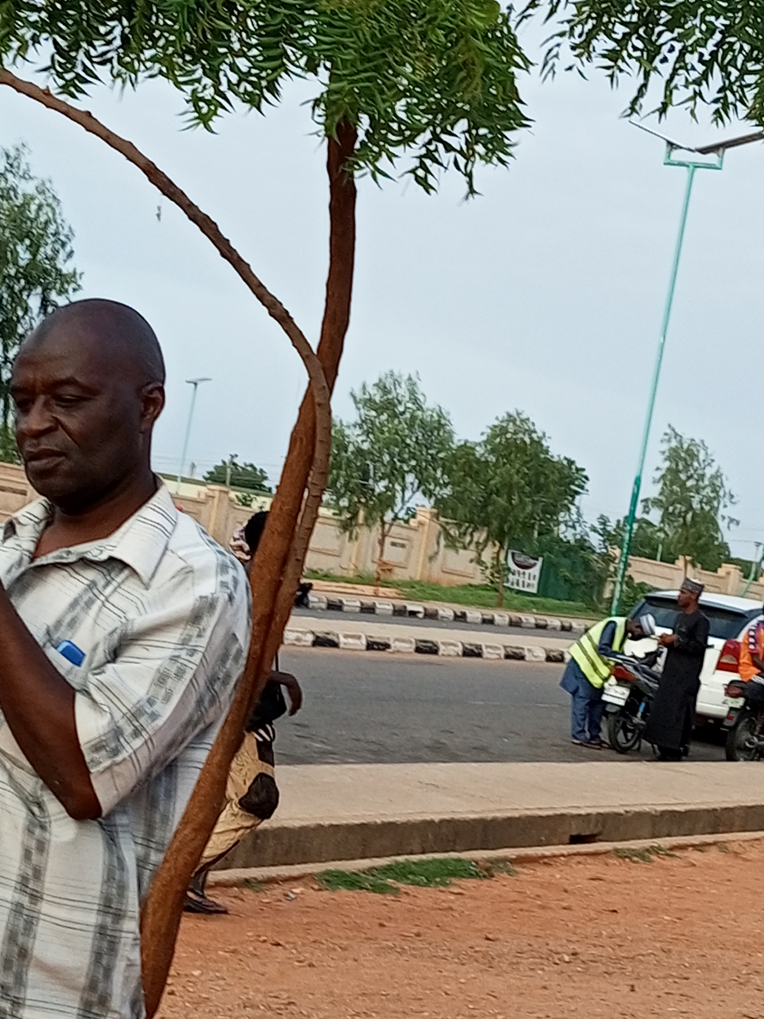 The image depicts an outdoor scene in Gombe, Nigeria. In the foreground, a man with dark skin and a shaved head is visible from the chest up, wearing a checkered collared shirt. He is standing near a tree with rough, brown bark and green leaves. The background shows a street with a white vehicle and two motorcycles. Several individuals are near the vehicles. One person is wearing a yellow safety vest and blue pants, appearing to interact with a motorcycle. Another person, dressed in dark clothing and a cap, stands beside them. Further down the street, a white wall with decorative elements lines the roadside, with trees and a cloudy sky above. The ground is a reddish-brown dirt.