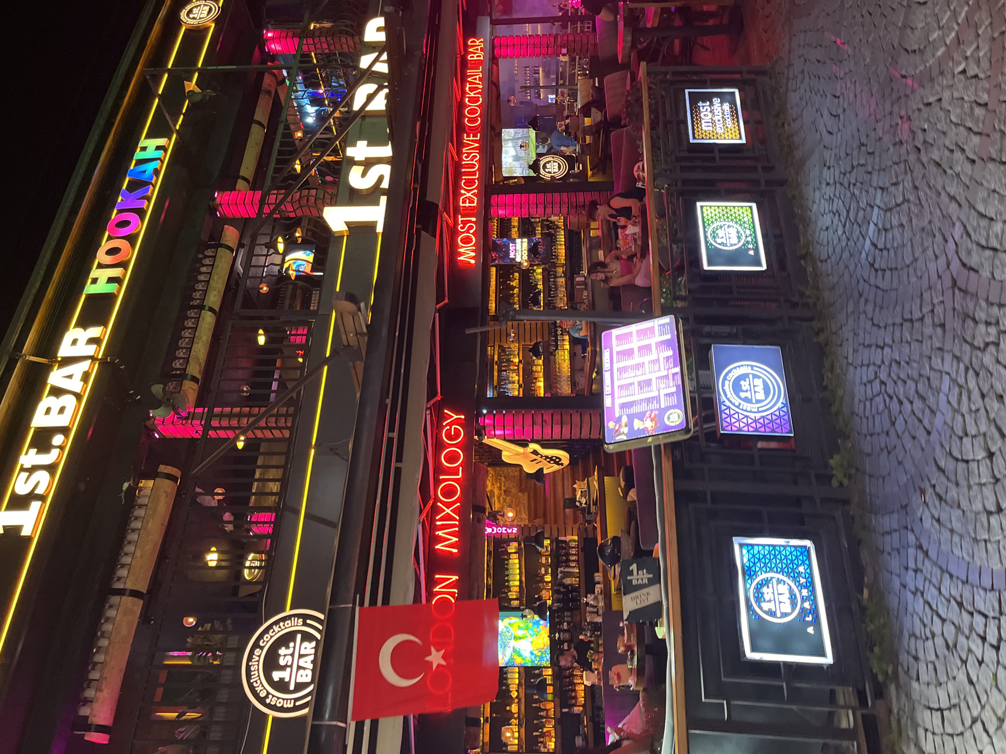 The image captures a vibrant bar or lounge at night, brightly illuminated by numerous neon signs. The scene suggests a lively nightlife atmosphere, likely in Alanya, Turkey, as indicated by a prominent Turkish flag hanging near the entrance and the text "ALANYA" on one of the signs.
The main establishment features a large, colorful sign on the upper left spelling out "1st. BAR HOOKAH." Below this, a red neon sign reads "LONDON MIXOLOGY," though part of it is obscured by the Turkish flag. A large, red neon sign running horizontally across the building prominently declares "MOST EXCLUSIVE COCKTAIL BAR."
Inside the bar, patrons are visible socializing and seated at tables. The interior is well-lit, showcasing a bar counter lined with bottles. Decorative elements include a white guitar-shaped cutout on an interior wall. The ground outside is paved with cobblestones.
Multiple smaller, illuminated display boards are arranged vertically to the right, advertising the establishment. These signs repeatedly feature "1st. BAR" and phrases like "most exclusive cocktails." One sign includes the word "cocktalle," likely a variant or misspelling of "cocktail." The overall impression is one of an energetic and popular spot for drinks and hookah.