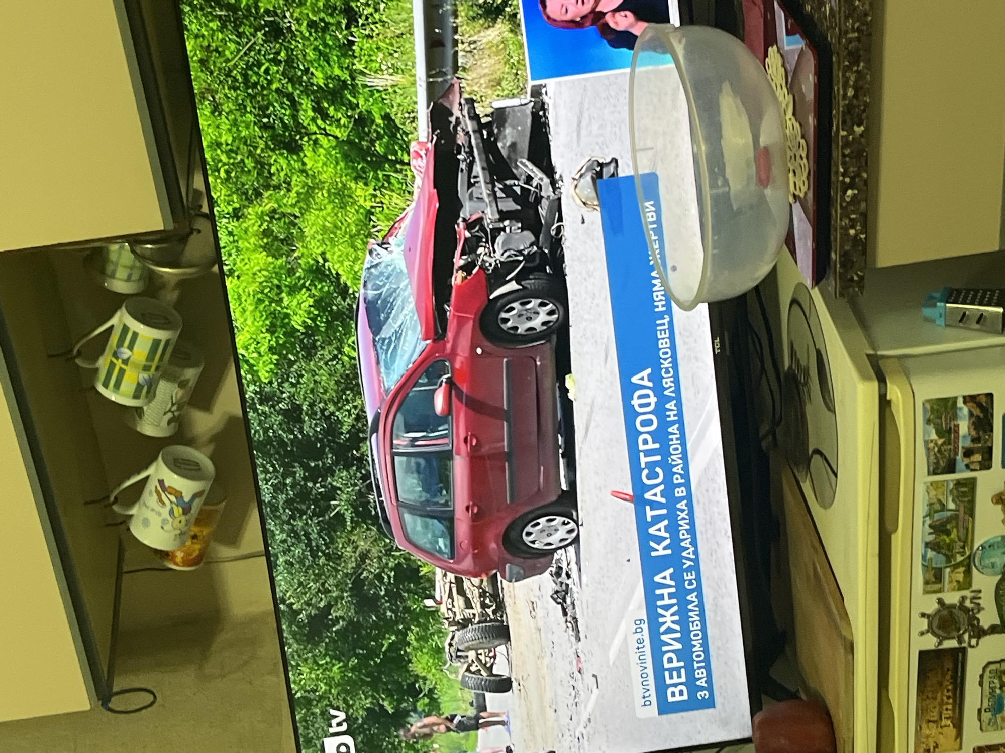 The image captures a widescreen television displaying a news report within a domestic kitchen or dining area, located in the city of Burgas, Bulgaria. The television screen presents an outdoor daylight scene showing the aftermath of a multi-vehicle collision on a paved road lined with dense green foliage. A heavily damaged red station wagon, possibly a Peugeot 307 SW, is prominently positioned on the right side of the road, facing left, with its front end severely crushed, windshield shattered, and the front driver's side tire appearing deflated. Further left, the chassis and rear axle of another significantly damaged vehicle are visible. Two shirtless male figures are standing on the left side of the road, observing the scene. A blue news banner at the bottom of the screen displays "btvnovinite.bg", the headline "ВЕРИЖНА КАТАСТРОФА" (CHAIN COLLISION), and the sub-headline "3 АВТОМОБИЛА СЕ УДАРИХА В РАЙОНА НА ЛЯСКОВЕЦ, НЯМА ЖЕРТВИ" (3 CARS CRASHED IN THE REGION OF LYASKOVETS, NO VICTIMS). A small picture-in-picture box on the far right shows a female news anchor speaking. Above the television, a light-colored shelf is mounted, from which several ceramic mugs hang from hooks; notable mugs include one with a cartoon character and another with a green and yellow striped pattern. The foreground features a dark, speckled countertop. On the right, a clear plastic bowl rests on a red square plate adorned with white decorative patterns. To the left, a wooden cutting board partially obscures a light-colored appliance, likely a refrigerator, whose side is decorated with various magnets, including landscapes, a ship's wheel, and text such as "ВЕЛИГРАД". A small silver box grater is also visible on the far left of the countertop. The television itself bears a "TCL" brand logo on its lower bezel.