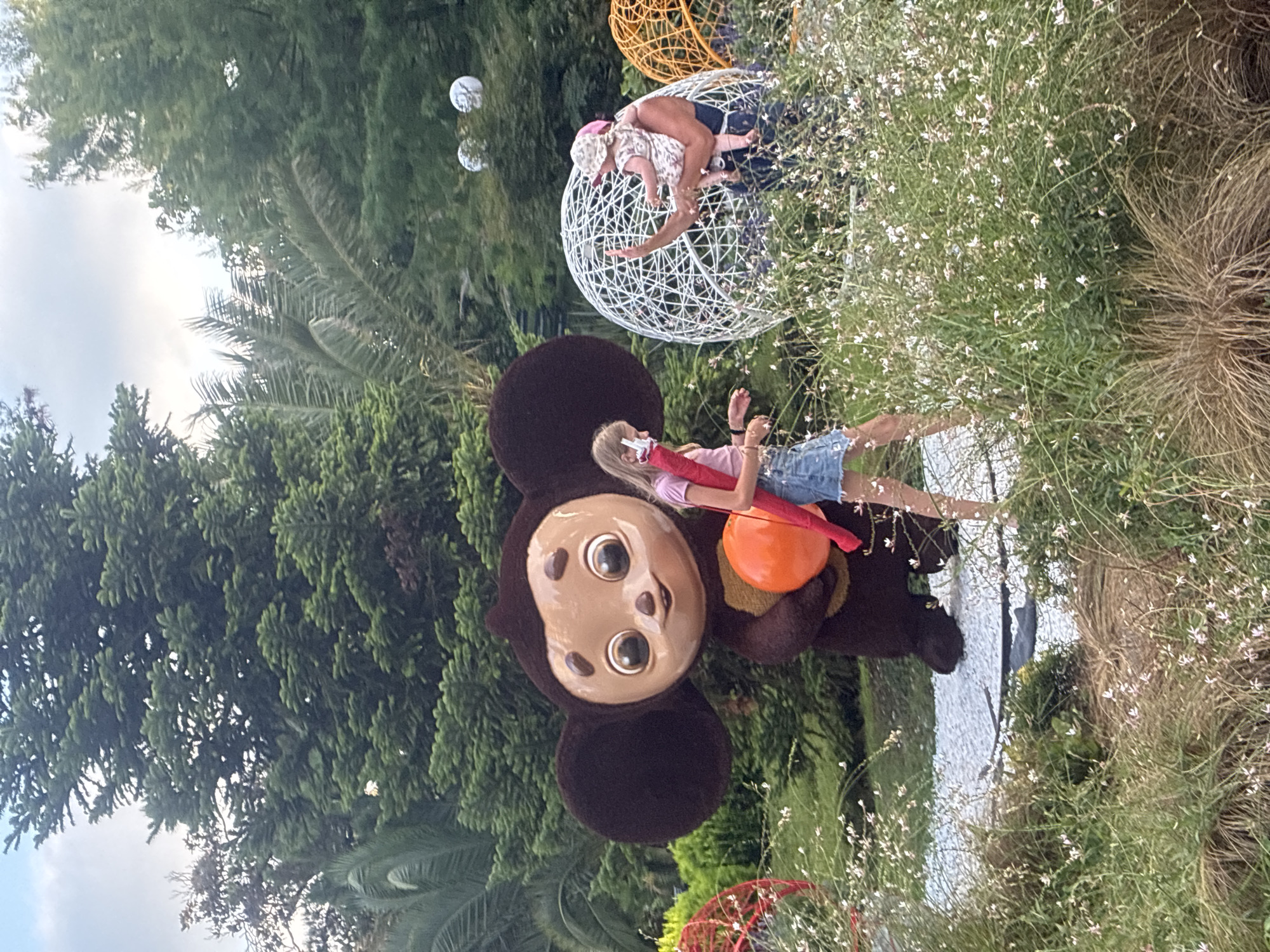 The image depicts an outdoor scene in a park-like setting in Sochi, Russia. In the foreground, a large character costume resembling Cheburashka, a popular Russian cartoon character, stands prominently. The character is brown with oversized ears and is holding a large orange sphere. To the right of the Cheburashka costume, a young girl with blonde hair, wearing a pink shirt and a denim skirt, is walking and holding a red umbrella. Further to the right, a person is holding a baby, who is wearing a hat, and interacting with a white spherical sculpture. The background features lush green trees and foliage, indicating a natural or landscaped environment. Several decorative spherical objects, some white and some orange, are visible throughout the scene, suggesting an artistic or recreational installation. The lighting suggests it is daytime, possibly late afternoon.