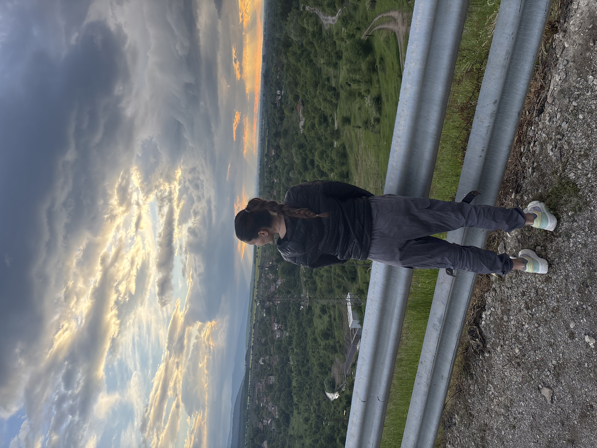 A young woman stands on what appears to be the edge of a bridge or high overlook in Ognyanovo, Bulgaria. She is leaning forward, looking down at the landscape below. She has dark hair in a braid, and is wearing a dark jacket and dark trousers with light-colored sneakers. A metal guardrail runs along the edge. Below the guardrail is a sloping hillside covered in green vegetation, leading down to a valley with scattered houses and buildings. The sky overhead is filled with dramatic clouds tinged with the orange glow of the setting sun.