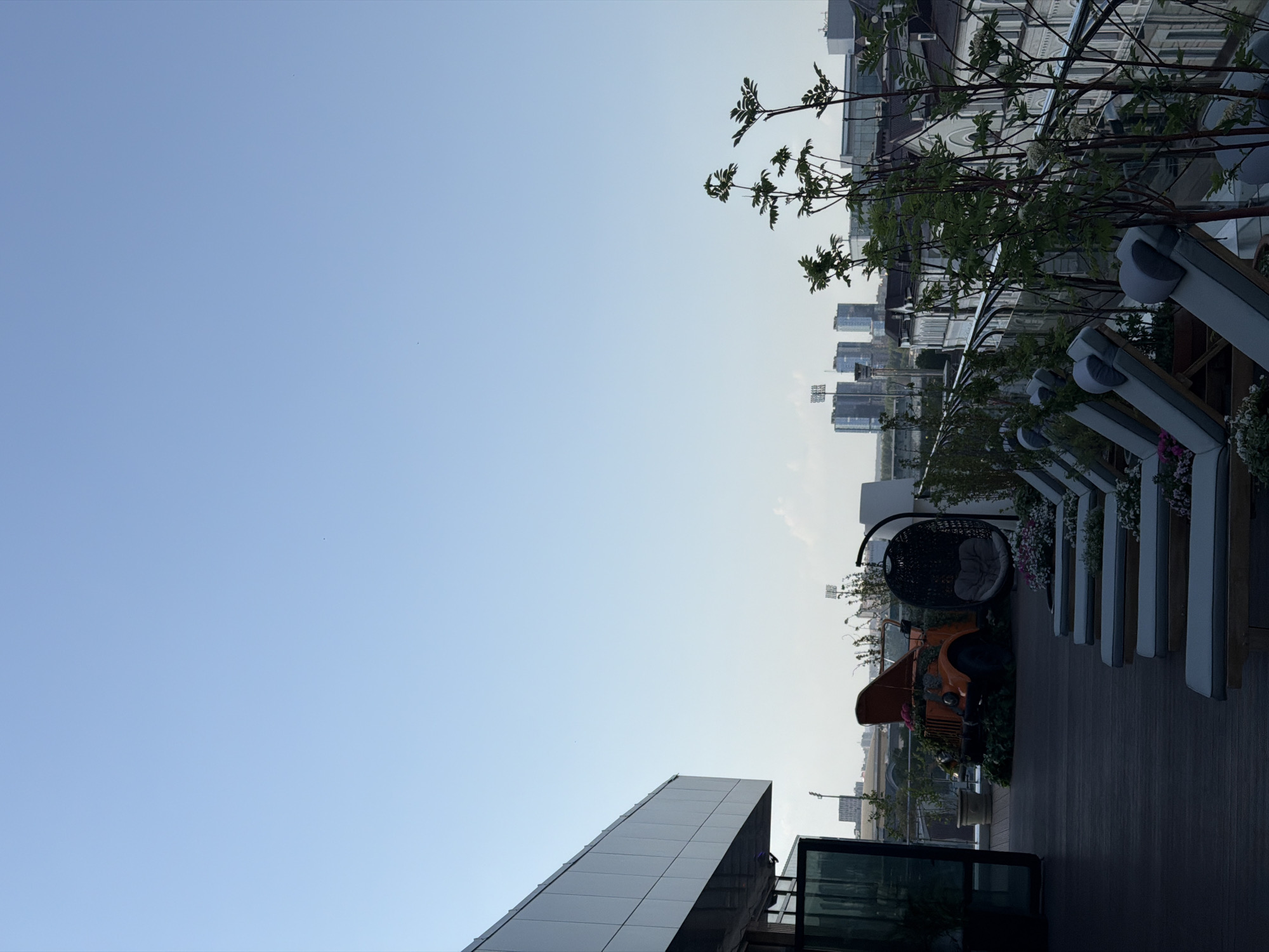 The image depicts an urban rooftop setting in Kazan, Russia, notable for its modern design and elevated perspective. The foreground presents a contemporary balcony or terrace arrangement, featuring cushioned benches with metal frames, integrated planters with flowers, a hanging wicker chair, and an orange vintage vehicle adorned with greenery. The background showcases a cityscape with several buildings in the distance, framed by trees and a clear sky. The overall scene suggests a stylized outdoor space designed for leisure and aesthetic appeal.
