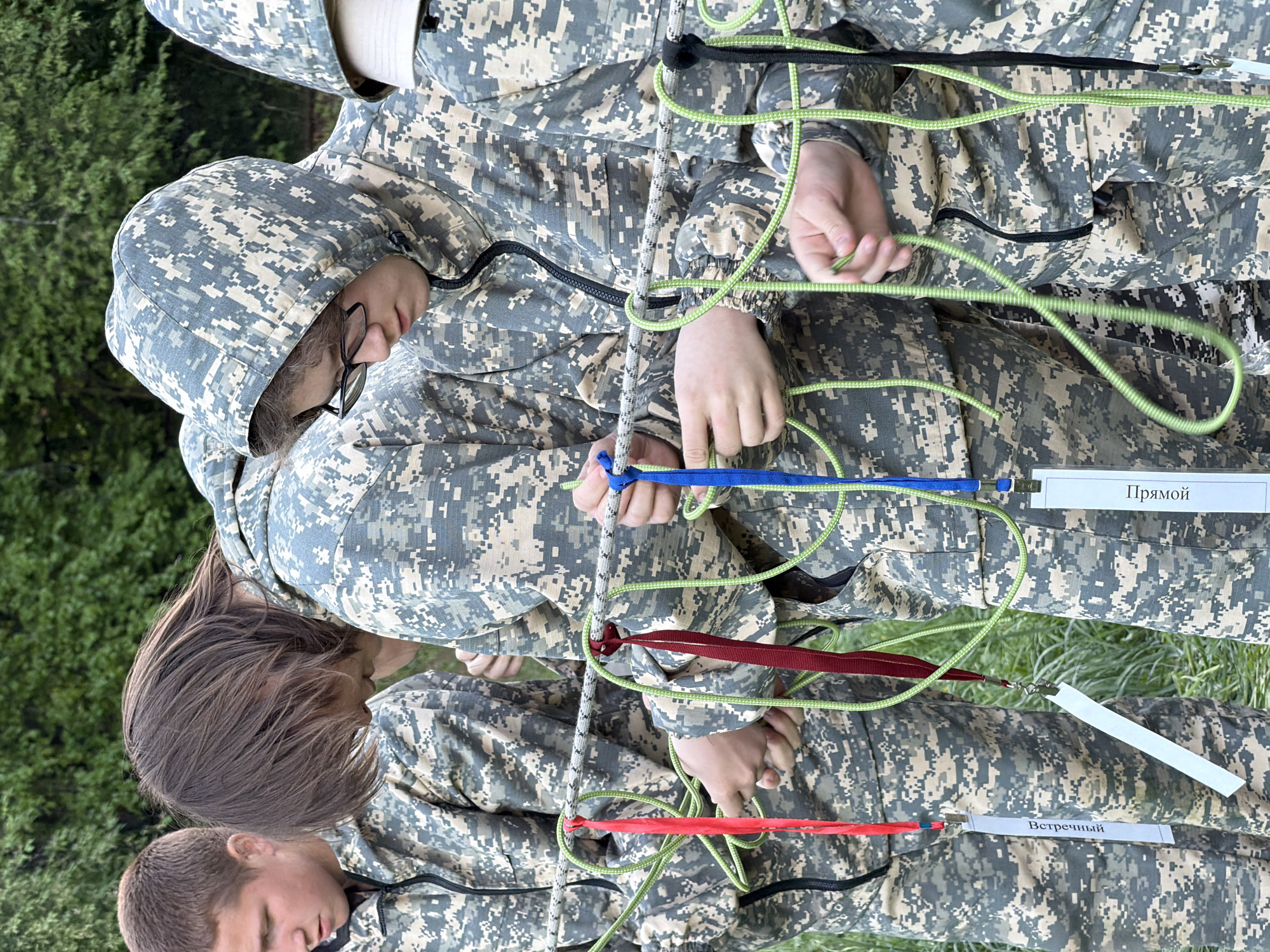 A group of four people in camouflage uniforms are participating in an activity involving ropes. The people are positioned close together, with one person wearing glasses and a hood pulled over their head. Each person is holding or interacting with the ropes, which are arranged in a complex pattern. Some of the ropes have colored tags attached to them, including blue and red. There are also white tags with text in Russian, visible on the right side of the image. The tag above shows the text "Прямой", the other one "Встречный". The background shows a green grassy area. No evidence proves the location is in Забегалово, Russia.