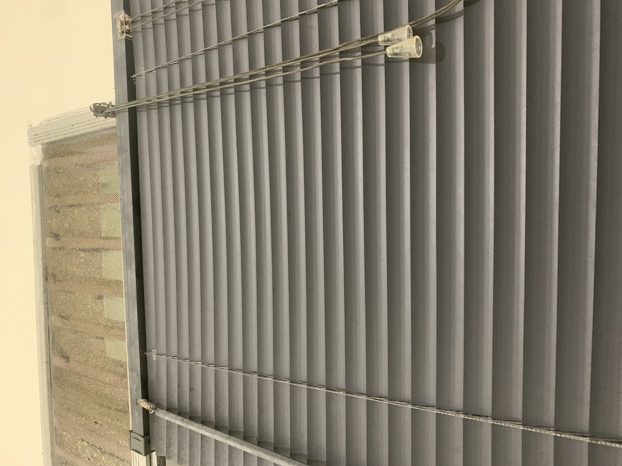 This media file captures a close-up view of a window with vertical blinds. The blinds are a dark grey color and appear to be made of fabric, with distinct vertical pleats. They are suspended by thin, horizontal cords that run across the width of the window.
Attached to these cords are two clear plastic clips, one of which is visible on the left side of the frame, and another is slightly to the right. These clips likely serve to secure the cords or possibly to hold the blinds in a particular position. The cords themselves have a slightly textured appearance.
To the left of the blinds, a portion of the window frame is visible. The frame is light grey and appears to be made of metal or a similar material. Within the window frame, there is a mesh screen, possibly a mosquito net, with a fine grid pattern. The screen has a slightly textured, woven appearance and some visible dust or debris.
Beyond the screen, the glass of the window is visible, though it is somewhat obscured by the screen and the angle of the photograph. A light-colored wall forms the background to the left of the window. The lighting suggests it is daytime, with natural light illuminating the scene from the left. There are no people or animals visible in the frame. The setting appears to be indoors, likely a room in a residential or commercial building. There is no discernible text in the image.