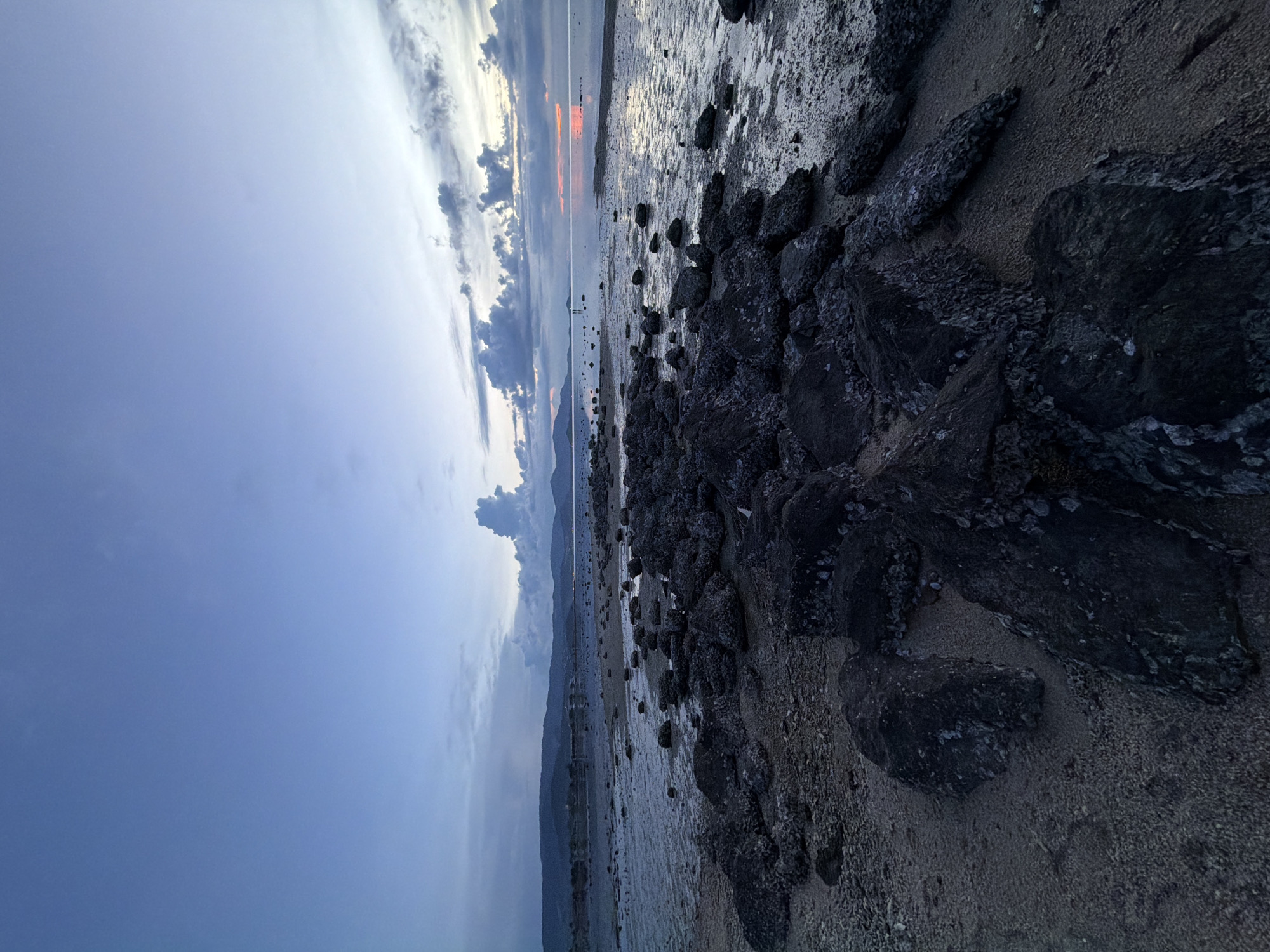 This image depicts a coastal scene at dusk in Baan Plai Laem, Thailand. The foreground is dominated by numerous dark, weathered rocks and pebbles scattered across a sandy and wet shore. Some of the rocks appear encrusted with barnacles or other marine life. The water's edge is visible, with shallow water reflecting the muted tones of the sky. In the middle ground, the calm sea stretches towards a distant, hazy shoreline with low-lying hills or mountains. The sky is a blend of deep blues and lighter grays, with scattered clouds that are illuminated by the setting sun, creating a soft, reddish glow on the horizon. The overall atmosphere is tranquil and serene. There are two small figures visible in the distance standing in the water.