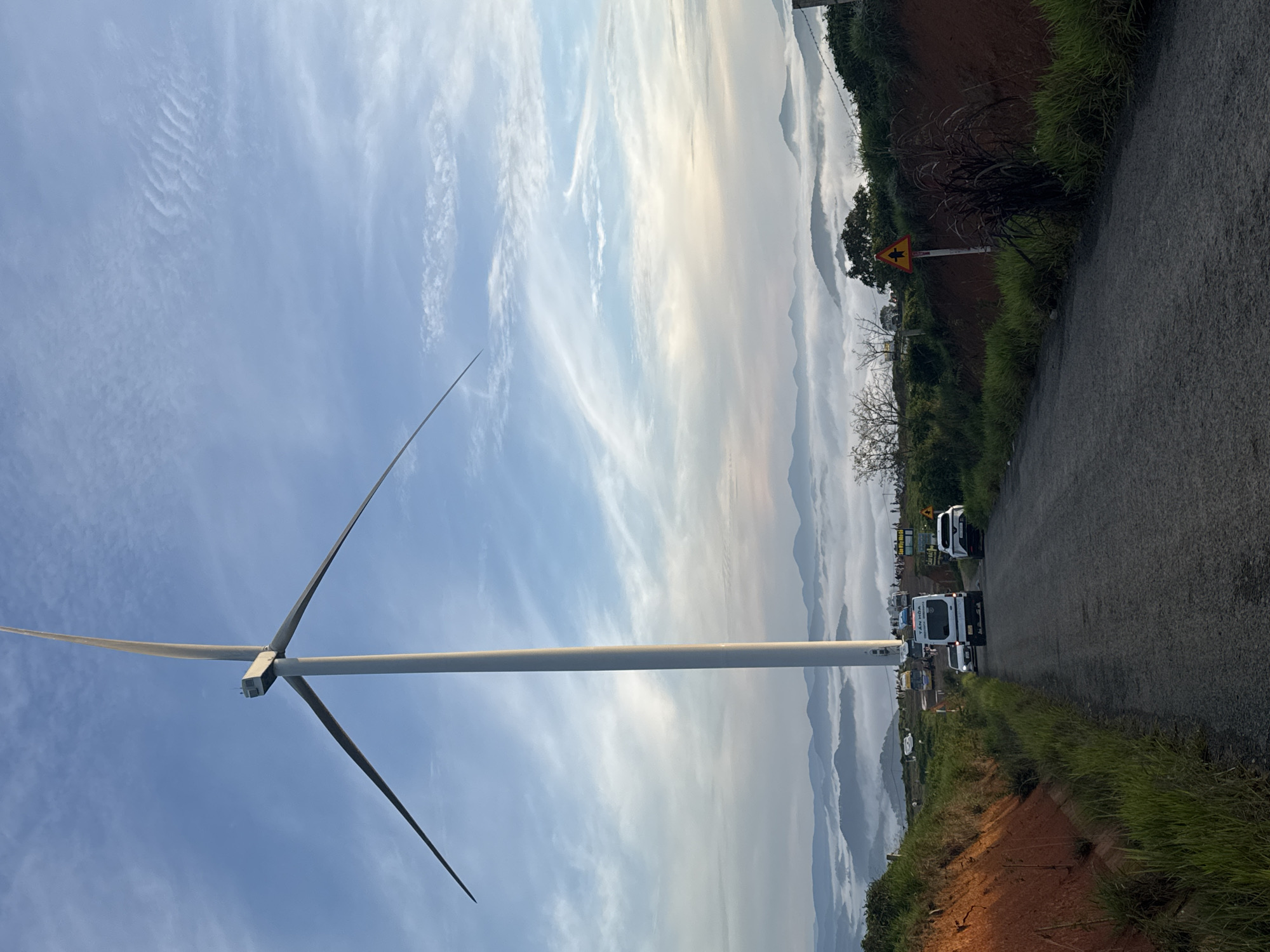 The image depicts a landscape in Phát Chi, Vietnam, dominated by a large wind turbine positioned prominently on the left side. The turbine, with its white tower and long blades, stands against a vast blue sky filled with wispy white clouds. In the background, rolling hills are enveloped in a thick layer of white fog, creating a serene, mountainous vista. A paved road curves through the scene, with several vehicles present. A white van, possibly a tour bus or shuttle, is visible on the road, along with other cars. The sides of the road are lined with green grass and reddish-brown soil. A triangular yellow road sign with a black symbol indicating a pedestrian crossing is visible on the right side of the road. A group of people are gathered near the base of the wind turbine, suggesting this is a tourist or scenic location. The overall atmosphere is tranquil, with the natural beauty of the landscape emphasized by the atmospheric fog and the presence of renewable energy infrastructure.