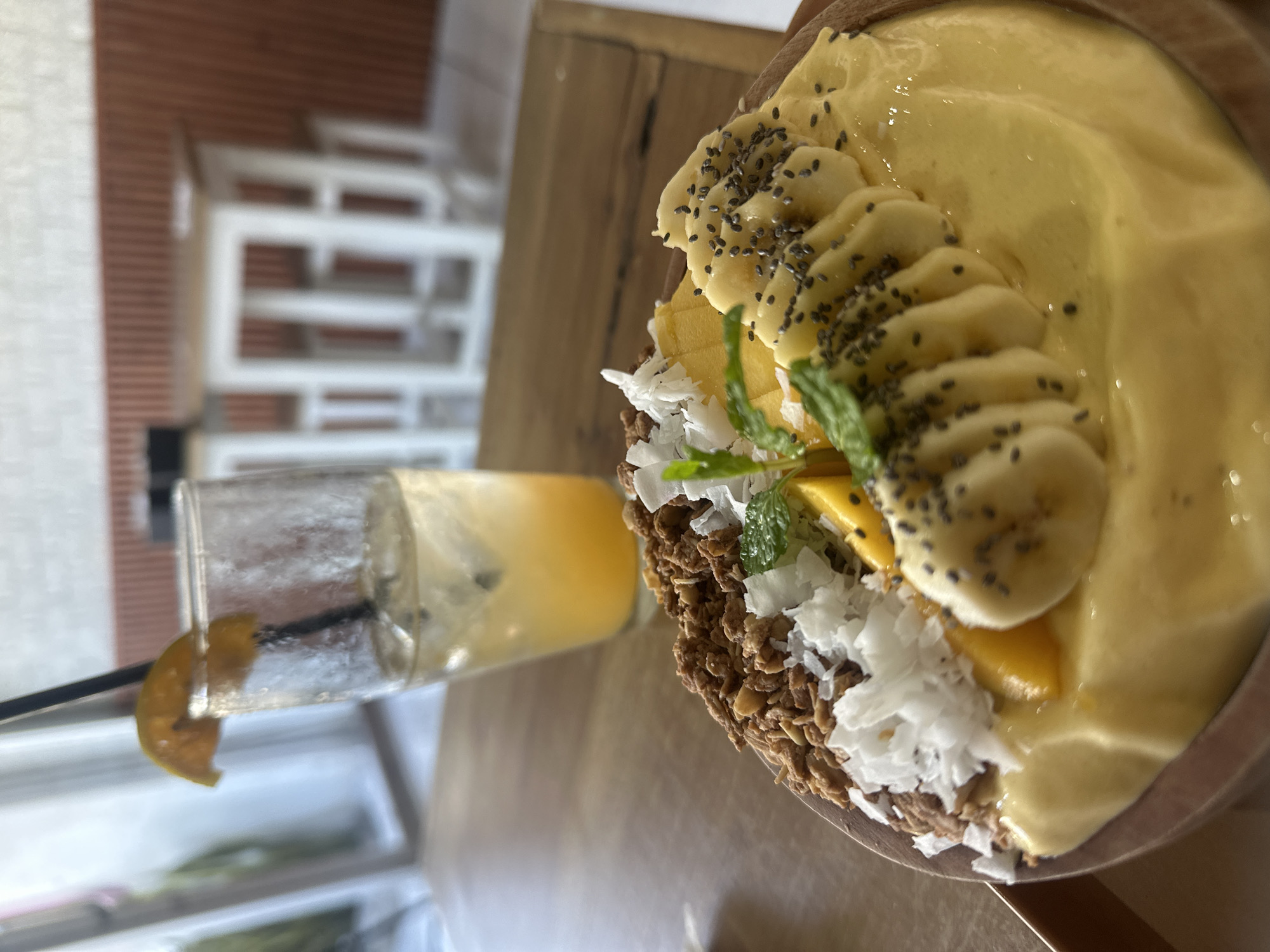 In Kuta Selatan, Indonesia, a close-up shot captures a breakfast bowl and a drink on a wooden table. The smoothie bowl, presented in a wooden dish, is filled with a vibrant yellow smoothie and topped with sliced bananas, chia seeds, shredded coconut, granola, and mint leaves. To the left of the bowl, a tall glass contains a light-colored beverage with ice, garnished with an orange slice and a black straw. The background features a blurred glimpse of a cafe setting with white chairs and tables, and a textured brown wall.