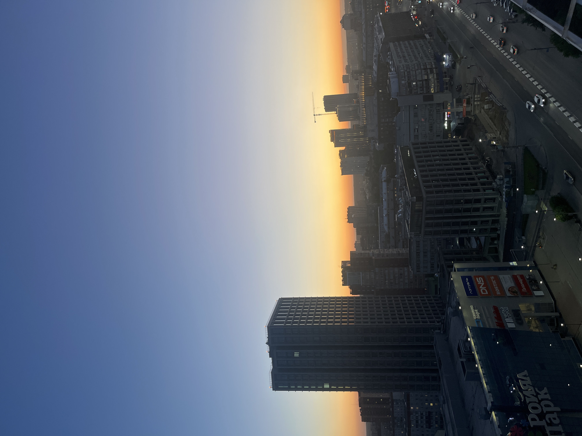 An aerial shot captures a cityscape at dusk, with a vast, gradient sky transitioning from deep blue at the top to warm orange and yellow hues near the horizon. In the foreground, the massive, dark facade of a shopping mall dominates the lower left, with illuminated signs for "Роял Парк," "Кино Стар," "DNS," "ROSTIC'S," "ЛЭТУАЛЬ," "ГАЛАМАРТ," and "ЧИТАЙ ГОРОД." Several cars are visible on a multi-lane road running parallel to the mall, with some streetlights illuminated. Beyond the mall, a dense cluster of high-rise buildings forms the urban skyline. A particularly tall, dark skyscraper with numerous windows stands prominently to the left of center. Further into the distance, more buildings of varying heights are visible, with a construction crane silhouetted against the colorful sky. The overall impression is one of a bustling urban environment at the close of day.