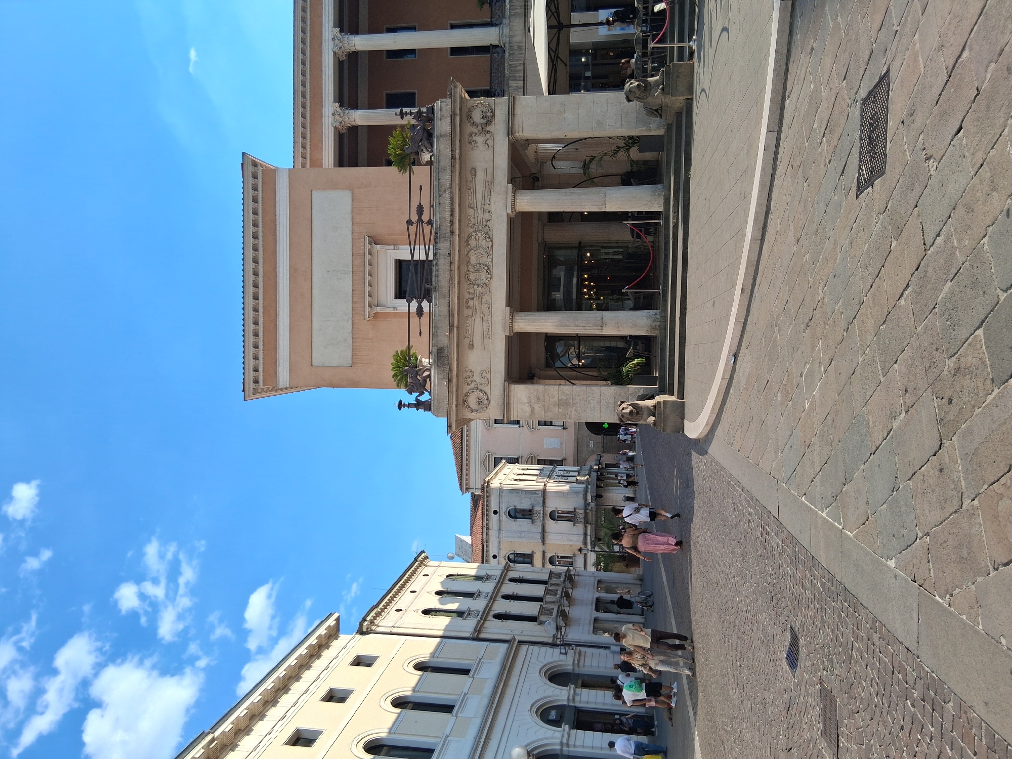 The image shows a sunlit piazza in Padua, Italy. In the foreground, the ground is paved with stone, transitioning from smaller cobblestones on the left to larger rectangular stones on the right. Several people are walking across the piazza.
On the left side of the image, a row of pale yellow buildings with arched windows and balconies lines the street. One building has an awning over its entrance. A few people are visible near the shops.
In the center and right of the image, a grand building with a large portico supported by Corinthian columns dominates the view. This building has a light pink facade and is adorned with decorative moldings and sculptures, including lion statues at the base of the portico. Red ropes guide visitors towards the entrance, and a cafe with outdoor seating and umbrellas is set up to the right of the building.
The sky is a vibrant blue with scattered white clouds. The overall scene depicts a lively urban square on a clear day.