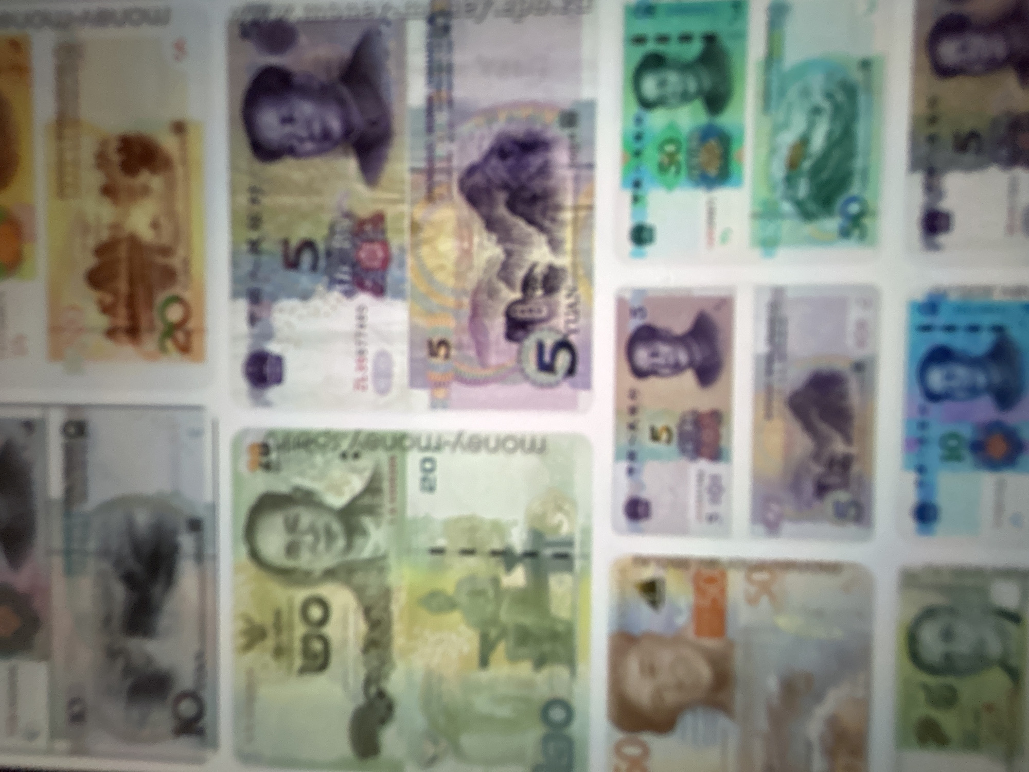 The media file is a photograph of multiple Chinese Yuan banknotes, arranged in a grid-like pattern. The banknotes are of various denominations, including 5, 10, 20, 50, and 100 Yuan. The image is somewhat blurry, making it difficult to discern fine details on the currency. Each banknote features portraits of people, likely historical figures or political leaders, which is common on currency. Some banknotes also display scenic imagery. The setting appears to be a flat surface, possibly a table or a wall, where the banknotes are displayed. There is no indication of a specific time of day, weather conditions, or explicit location cues beyond the context of Gelendzhik, Russia, which is provided separately. The overall impression is of a collection or display of Chinese currency. Text visible on the banknotes includes the denomination numbers (e.g., "5", "10", "20", "50", "100") and the word "Yuan". Some other characters and symbols are present but are too blurred to be clearly identified. The emotions conveyed are neutral, as the image focuses on the objects themselves.