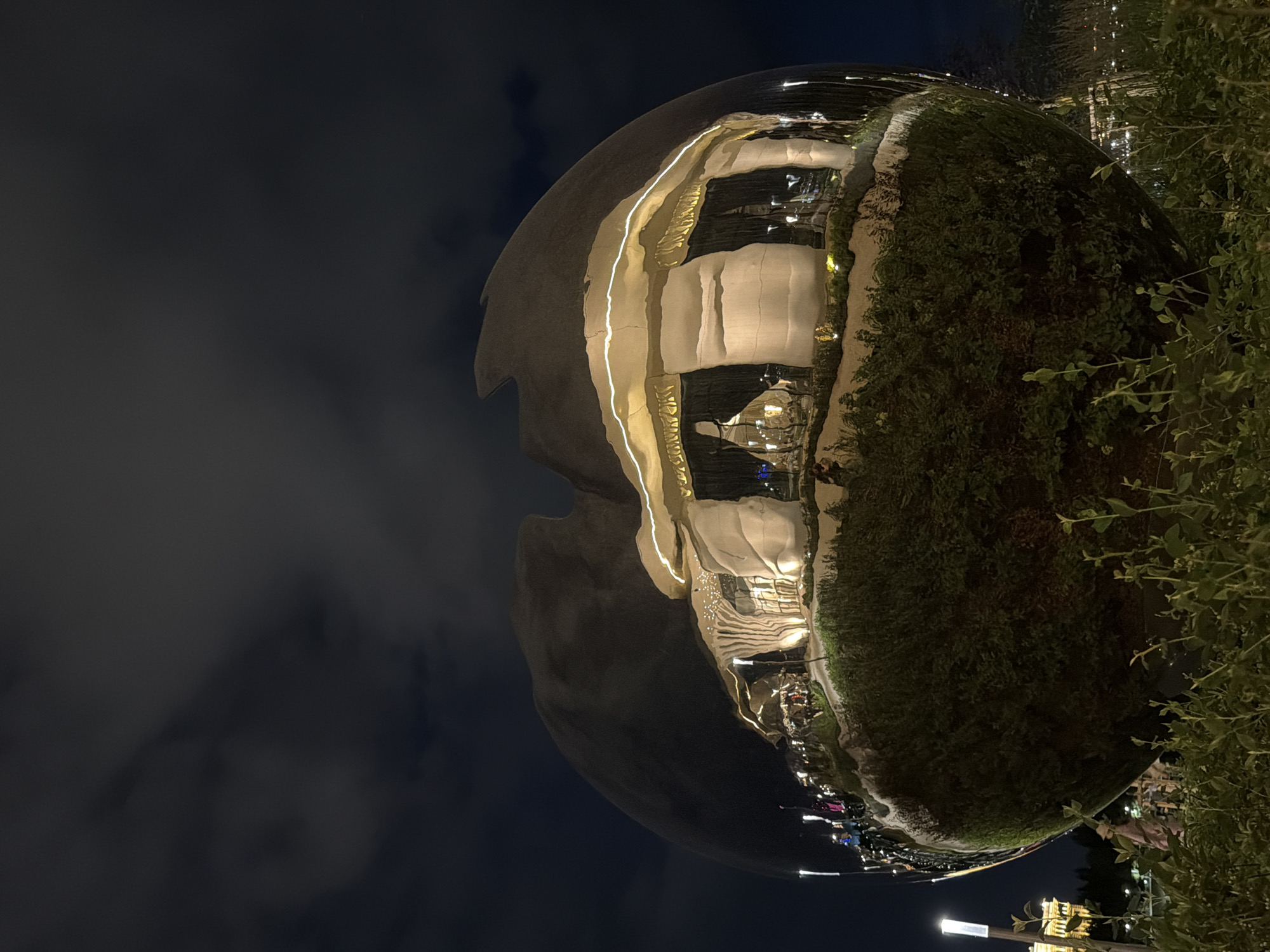 The image shows a large, highly reflective, spherical sculpture located in Astana, Kazakhstan, as indicated by the distinctive architecture reflected on its surface. The sculpture is set against a dark night sky with visible clouds. The reflection on the sphere depicts a modern building with illuminated windows, organic, mushroom-like architectural elements, and string lights. The base of the sculpture is surrounded by lush green bushes and foliage, with some taller plants in the foreground. In the background, a street lamp with a white light is visible, and further back, a softly lit building with a classical facade can be seen. The overall scene suggests an outdoor public space at night.