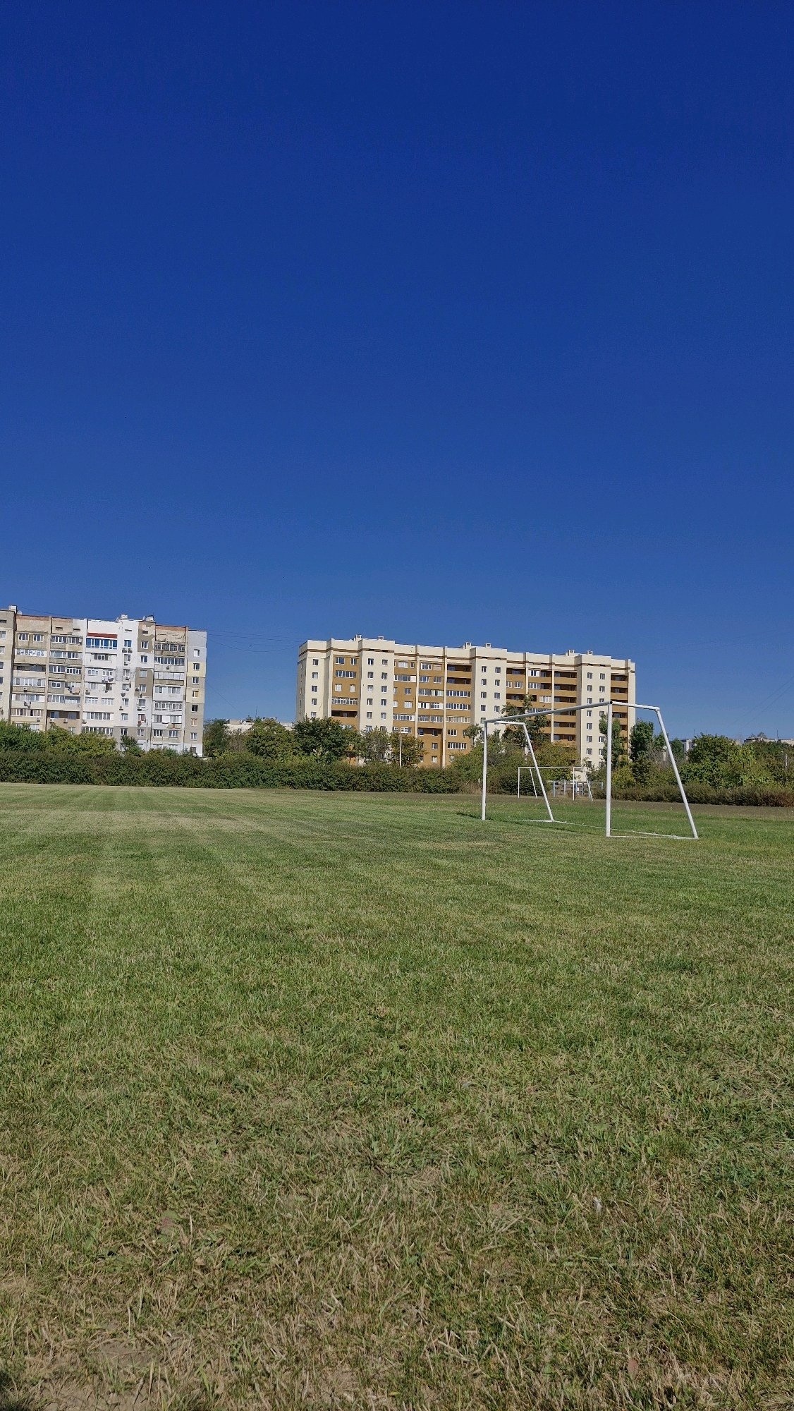 The image depicts a bright, clear day in Serhiivka, Ukraine. The foreground is dominated by a vast, well-maintained green lawn, likely part of a sports field or park. In the mid-ground, a white soccer goal is prominently positioned on the right side. Behind the goal and extending across the frame are several multi-story apartment buildings, characteristic of Soviet-era architecture. These buildings vary in color, with one on the left appearing mostly white and gray, and a larger one on the right featuring beige and yellow tones. A dense line of trees and shrubs separates the grassy area from the apartment buildings. The sky is a vibrant, cloudless blue, occupying the upper portion of the image. The overall scene suggests a typical urban residential area with recreational facilities.