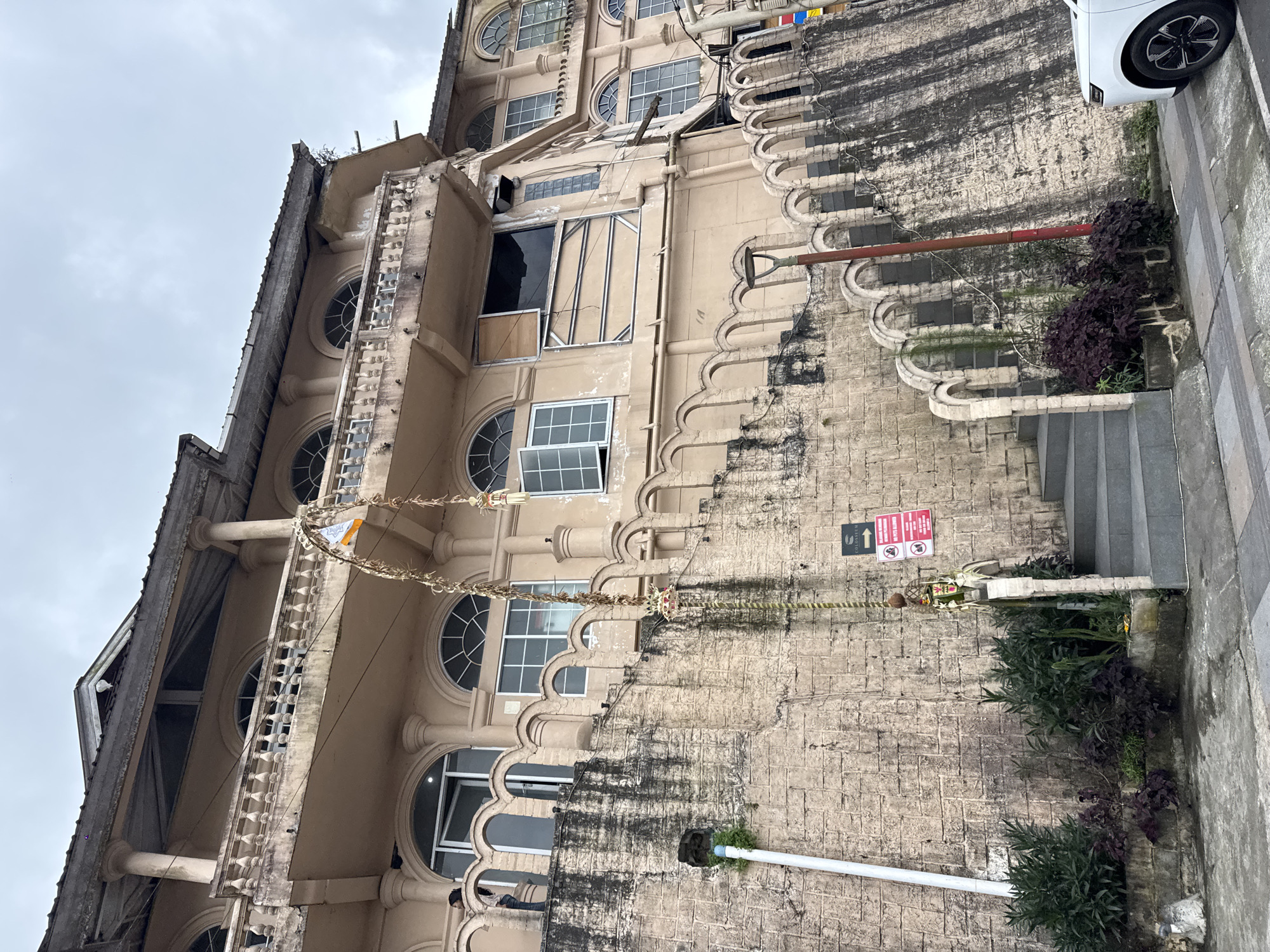 In Kintamani, Indonesia, a multi-story tan building with a faded aesthetic is observed, characterized by arched windows, a prominent balcony with decorative railings, and evidence of structural wear. A large stone wall ascends to the building's lower levels, featuring an arched design along its upper edge. A set of concrete stairs leads up the wall, flanked by greenery and a red lamppost. A white car is partially visible on the right side of the frame. A decorative Balinese offering, suspended from an archway, hangs in the center of the image. Two signs are affixed to the stone wall near the stairs, one indicating "COLOSSEUM" with an arrow, and another displaying a symbol and text in red and white, likely a warning or information sign. The sky is overcast with clouds.