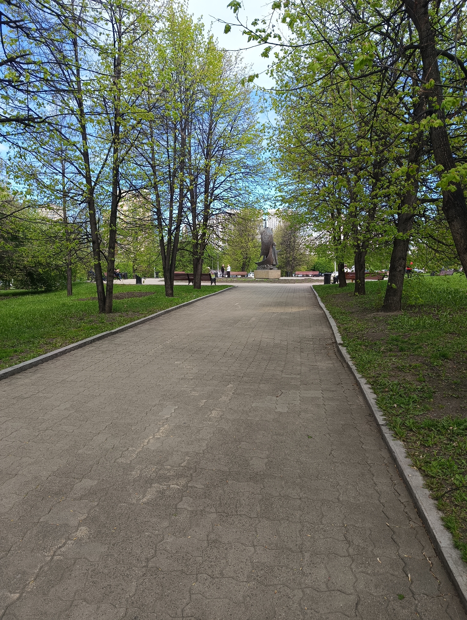 The image depicts a park pathway in Yekaterinburg, Russia, leading toward a statue of a ship. The pathway is paved with interlocking gray stones and flanked by green grass and trees with budding leaves. Benches are situated along the pathway, with a few people visible in the distance, either sitting or walking. Buildings are visible in the background beyond the trees. The sky is blue with scattered clouds.