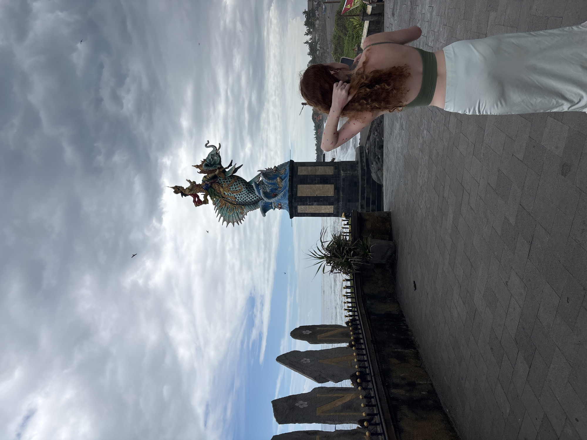 A full shot, in daylight, shows a woman with auburn curly hair, wearing an olive-green top and light-green skirt, standing near a colorful statue. The statue depicts a figure riding a sea creature with a dragon's head. It stands on a dark, multi-tiered pedestal, situated along a paved walkway.
Behind the statue is a view of the ocean and the Mengwi coastline, under a sky filled with heavy, mottled clouds.
The camera angle is slightly low, creating a tilted perspective that makes the subjects appear in a diagonal orientation. In the foreground are stone structures with decorative elements, indicating a public walkway or viewing area.