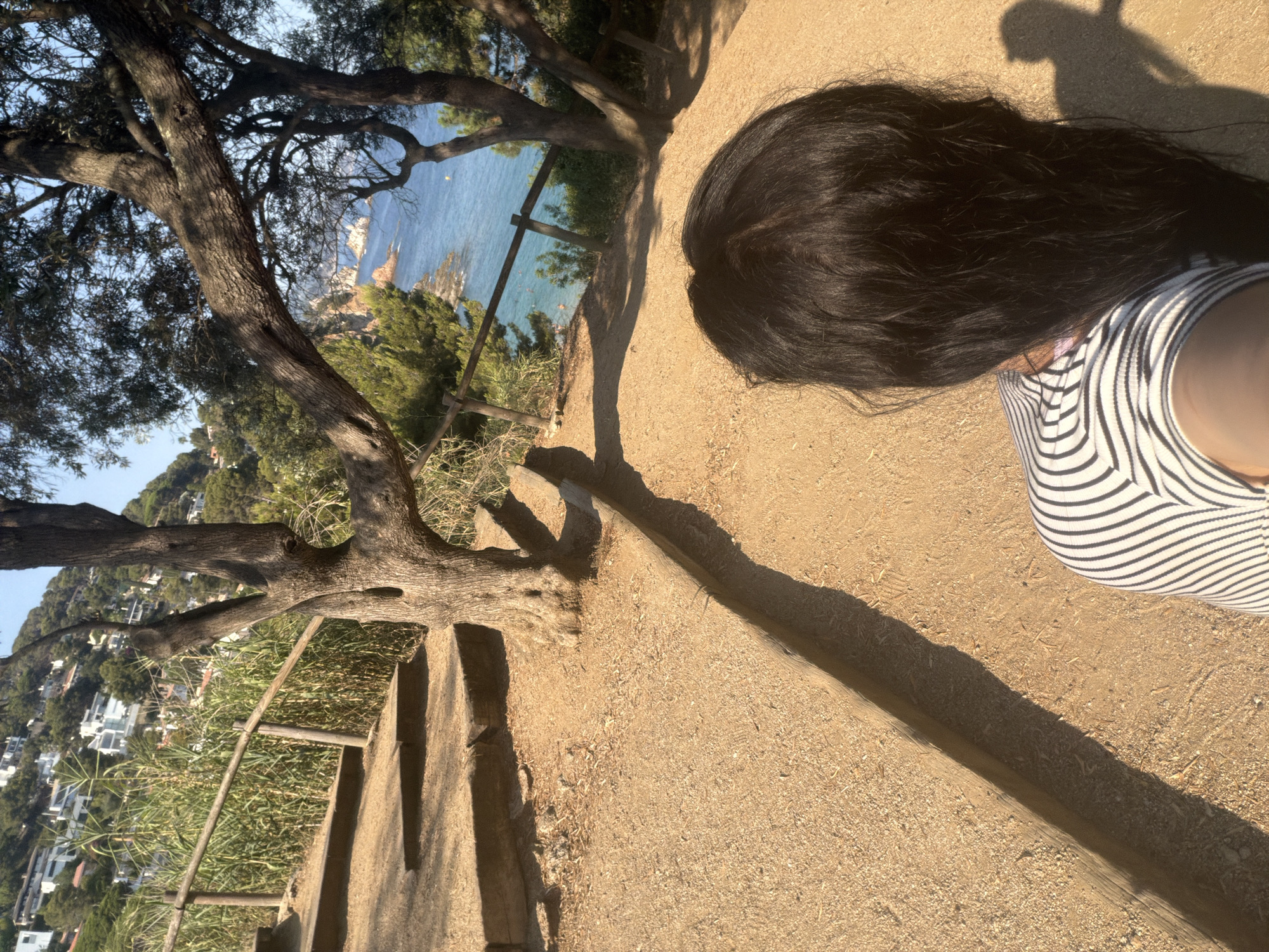 The image captures a first-person perspective of a person with long, dark hair wearing a black and white striped top. They are standing on a gravel path with wooden steps ascending to the left, bordered by a wooden railing and lush green vegetation. In the background, the deep blue Mediterranean Sea is visible, with rocky outcrops and distant buildings nestled on a hillside. The scene appears to be a scenic coastal trail, likely in or around Blanes, Spain, given the distinctive cliffside architecture and the characteristic Mediterranean vegetation and coastline. The lighting suggests it is daytime, possibly late afternoon, casting long shadows from the trees and the person.