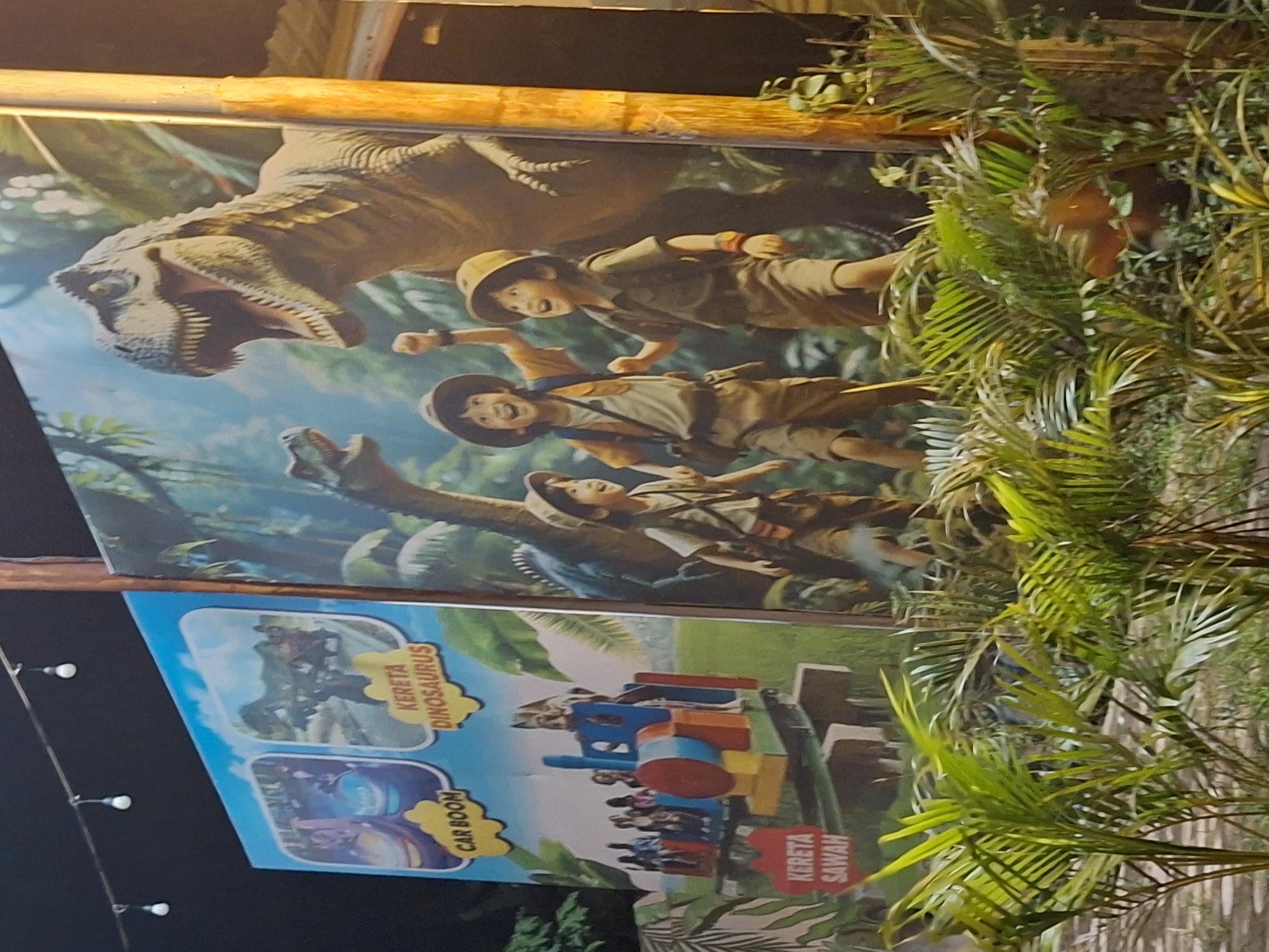 A nighttime outdoor shot captures a billboard featuring a dinosaur-themed advertisement. The billboard shows a Tyrannosaurus Rex and three people dressed in explorer outfits. Below this scene are smaller panels displaying "Kereta Dinosaurus" (Dinosaur Train), "Car Boom," and "Kereta Sawah" (Paddy Field Train) attractions. The billboard is partly obscured by lush green foliage, particularly palm fronds. In the background, a row of lights indicates the location is likely in an urban area. Given the references on the billboard, it is likely that this image was taken in Lhokseumawe, Indonesia, at an amusement park or similar tourist destination.