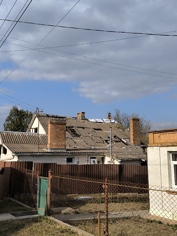 The image depicts a residential scene in Vinnytsia, Ukraine, under a partially cloudy blue sky crisscrossed by multiple utility lines. In the foreground, a rusty brown chain-link fence with a small green metal gate encloses an uncultivated plot of land. Behind this, a dark brown corrugated metal fence partially obscures the lower section of a two-story house. The primary subject is this house, which features a damaged grey corrugated roof. Numerous sections of the roof material are broken, missing, or displaced, revealing exposed structural elements beneath. Two brick chimneys are visible on the roof. A person, dressed in dark clothing and a cap, is positioned on the roof towards the right side, actively working on a section of the damage, possibly installing or adjusting light-colored sheeting. The house walls are light-colored, with visible windows on the lower level. To the far right, part of an adjacent building with a lighter grey facade and brick accents is visible. Trees are present in the background on the left. The scene is illuminated by natural daylight.