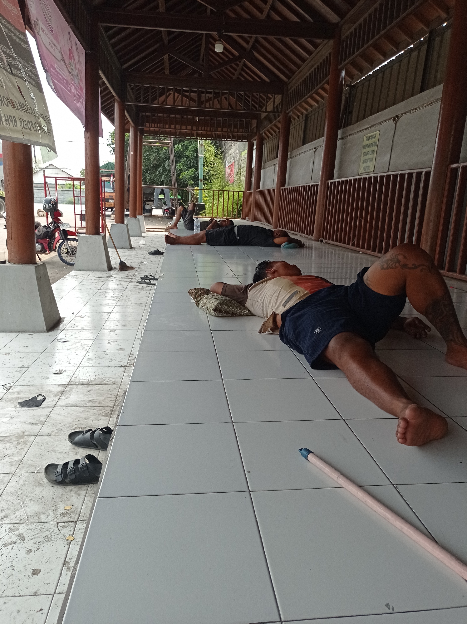 The image depicts a covered outdoor area with a tiled floor. Several people are lying down and sleeping on the floor. In the foreground, a man with tattoos on his leg is sleeping on his back, wearing a t-shirt and shorts. To his left, another person is sleeping on their side. Further back, two more individuals are visible sleeping. Various pairs of sandals and flip-flops are scattered on the floor. A red motorcycle is parked to the left of the frame, and a broom leans against a pillar. Banners are displayed on the left side of the structure, and a wooden fence runs along the right side. The environment appears to be a public waiting or resting area in Kramat, Indonesia, likely a transit point or a place of worship, given the covered structure and the presence of sleeping individuals.