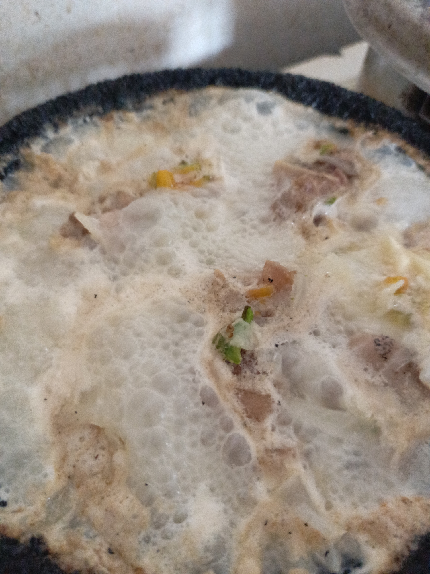 A close-up shot depicts a partially cooked omelet in a dark-rimmed pan. The omelet appears light in color with visible bubbles, indicating it is still in the process of cooking. Embedded within the omelet are small pieces of chopped vegetables, including what appear to be carrots and green onions, along with scattered bits of meat. The location, Juan Griego, Venezuela, is not discernible from the image itself.