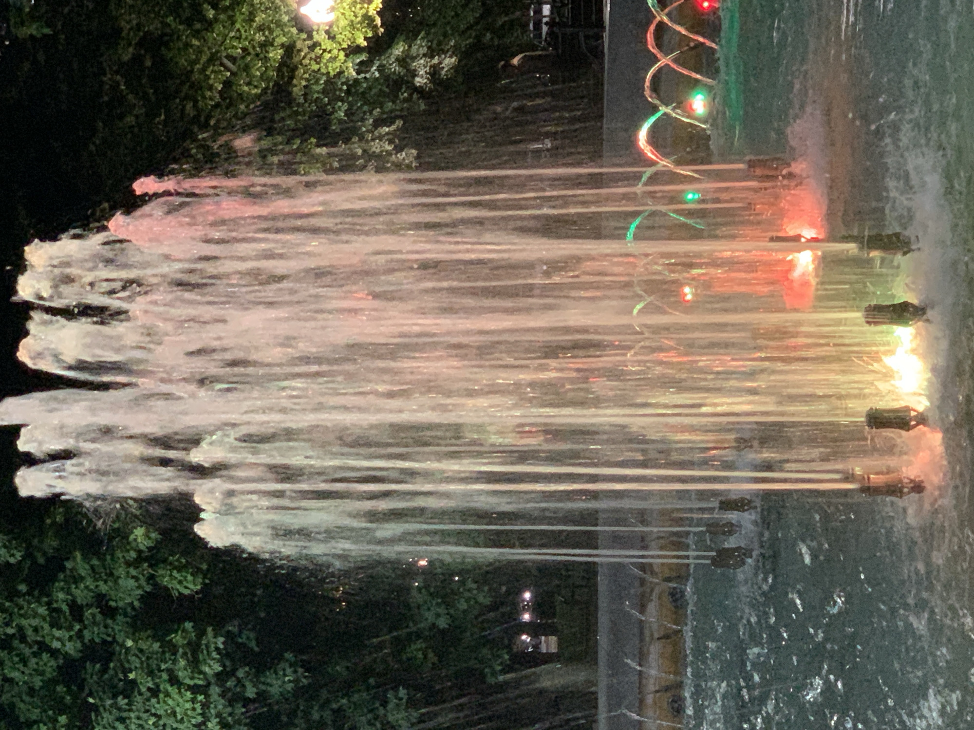 A nighttime shot shows a multi-tiered fountain in the foreground. The fountain jets are illuminated with red and green lights, creating a colorful display against the dark background of trees and night sky. In the background, a person can be seen sitting on a bench, and a vehicle is parked nearby. The water in the fountain is turbulent and appears to be splashing around the base of the jets. The overall atmosphere is one of tranquility and beauty, with the vibrant lights of the fountain contrasting with the dark surroundings.