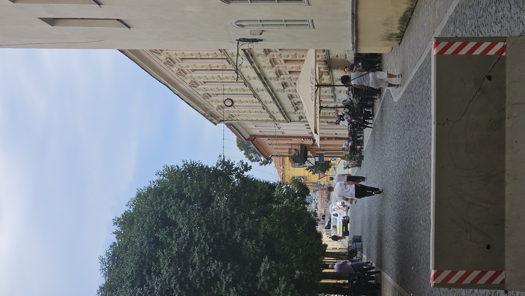 The image depicts a street scene in the city of Prešov, Slovakia. The foreground features a concrete barrier with red and white diagonal stripes on its ends. A cobblestone street extends into the background, lined on the right by a cream-colored building with ornate architectural details and arched windows. Tables and umbrellas for outdoor dining are set up along the sidewalk in front of this building. To the left, a large tree with dense foliage casts shadows on the street. Several people are visible walking along the street and seated at outdoor cafes. The sky is overcast. In the distance, more buildings and trees are visible, along with traffic and street signs. The overall impression is that of a lively European city street on a cloudy day.
