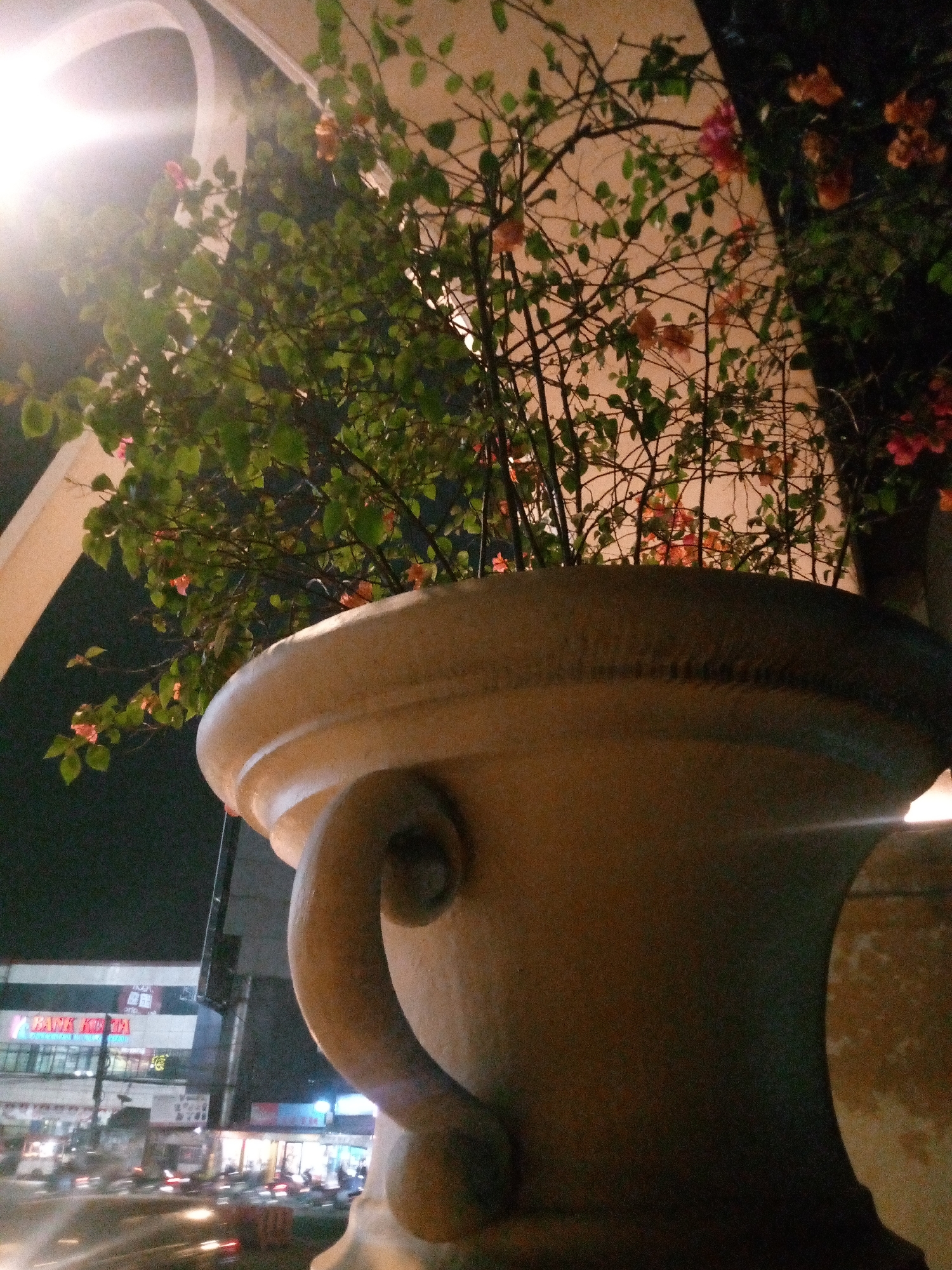The image shows a large, ornamental pot with plants and flowers in front of a brightly lit building. The pot is made of a light-colored material, possibly concrete or ceramic, and has a decorative handle. The plants are green and leafy, with some small pink and orange flowers visible. In the background, there is a building with signs that read "BANK KOTA" and other text in Indonesian. There are also streetlights and what appear to be vehicles on a street in front of the building. The overall scene suggests an outdoor setting at night, possibly in a commercial area. The location is identified as Bitung Jaya, Indonesia.