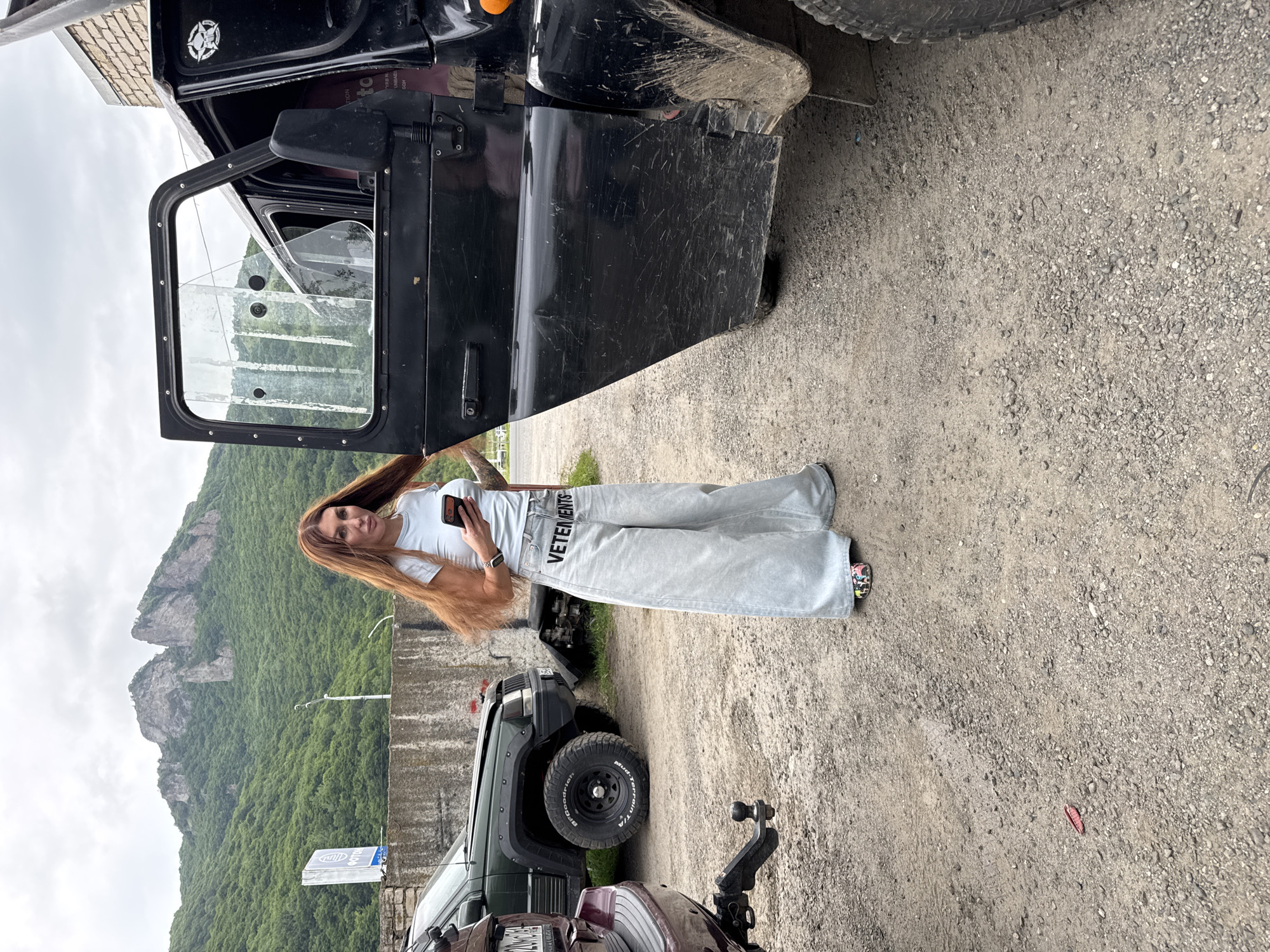 In the city of Karachayevsk, Russia, a woman with long, red hair stands next to an open black vehicle door. She is wearing a light blue t-shirt and light-wash, wide-leg jeans with "VETEMENTS" printed in black on the left leg. She holds a smartphone in her right hand. The vehicle appears to be a rugged, off-road type, with dirt and scuffs visible on its exterior. To the left of the black vehicle, another dark-colored SUV is parked. In the background, a rocky, tree-covered mountain range rises under a cloudy sky. The ground is covered with gravel and dirt, suggesting an unpaved area. A red object, possibly a piece of litter, is visible on the ground in the lower left quadrant of the image.
