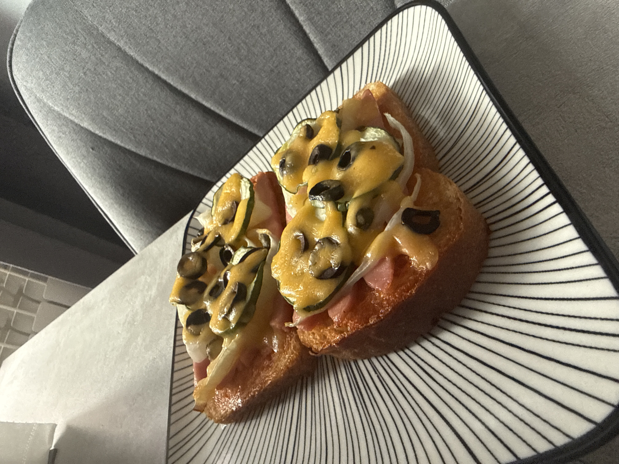Two open-faced sandwiches are presented on a white plate with black radiating lines. The sandwiches are made with toasted bread topped with ham, cheese, sliced cucumbers, white onions, and black olives. The plate is situated on a textured gray surface, possibly a table, with a portion of a gray upholstered chair visible in the background. There are no people or animals in the image, and no specific landmarks to determine the location in Verkhnezeysk, Russia. The lighting suggests an indoor setting, possibly a home or restaurant.