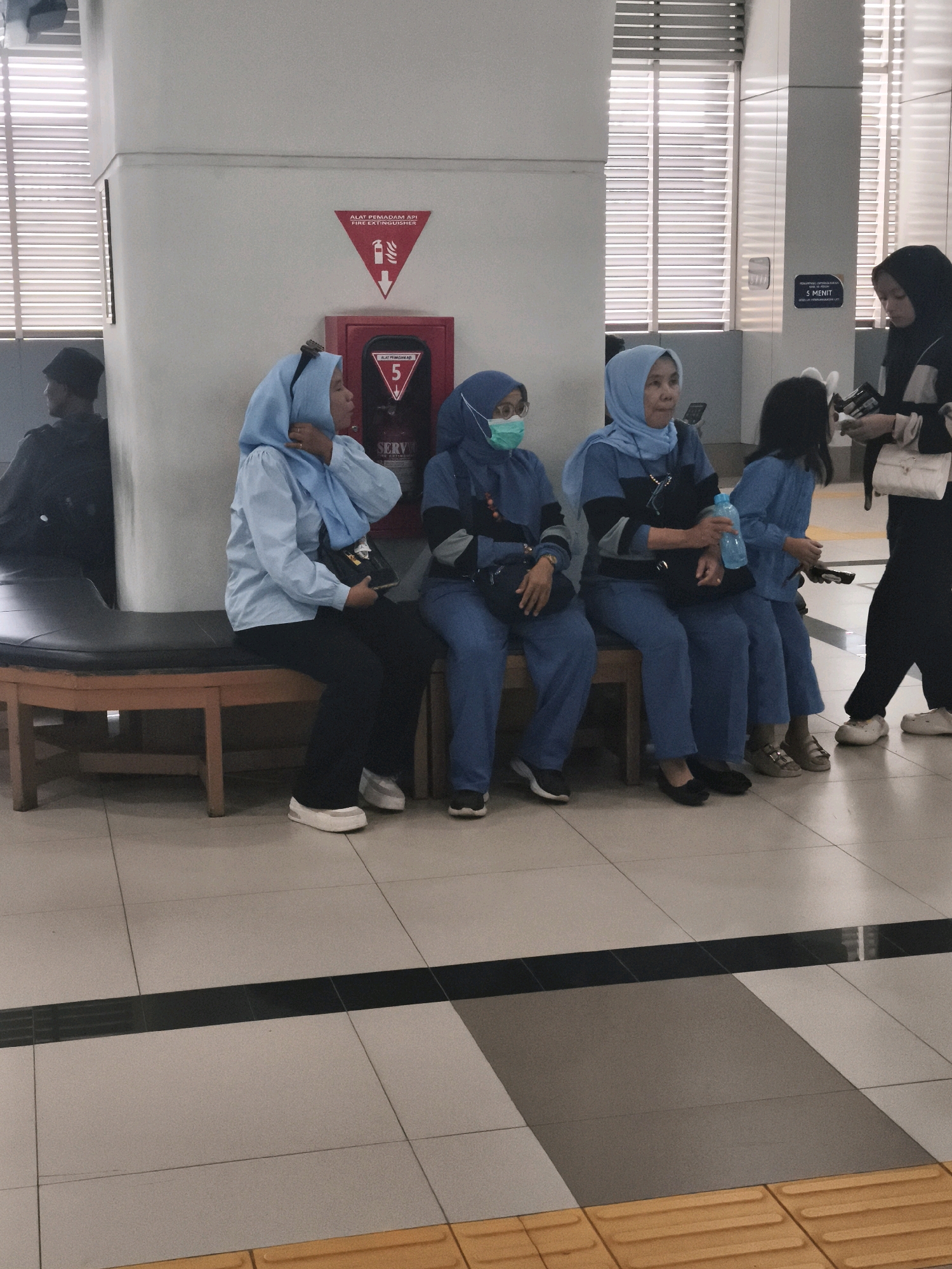 A group of people, mostly women wearing blue uniforms and headscarves, are sitting on a bench in what appears to be a waiting area, possibly a train station or bus terminal. The environment is indoors, with white walls and tiled flooring. A fire extinguisher is visible on the wall behind the seated individuals. One woman on the left is wearing a green surgical mask. Another woman in the center is holding a blue water bottle. To the right, a woman in black is standing and interacting with someone out of frame. A child in a blue outfit is also seated on the bench. The overall impression is one of waiting or transit. The location is identified as Kelurahan 16 Ilir, Kec. Ilir Timur I, Indonesia.