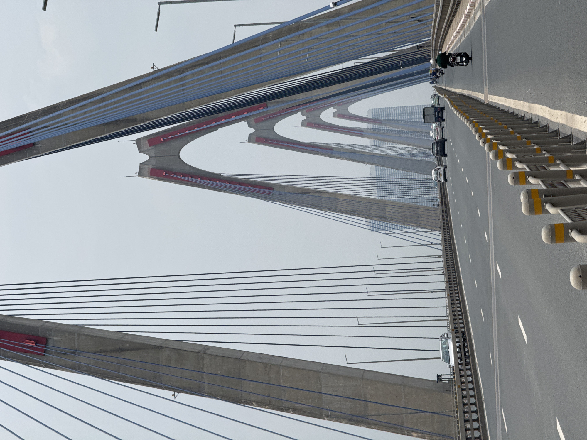 The image depicts the Nhat Tan Bridge, a cable-stayed bridge spanning the Red River in Hanoi, Vietnam. The perspective is from the road deck, looking towards the middle of the bridge. Several imposing concrete pylons, painted with red accents, dominate the scene, supporting numerous blue and black cables that stretch across the sky. The bridge deck is a multi-lane road with traffic, including cars, trucks, and motorbikes. In the distance, modern high-rise buildings are visible through the hazy sky. The road is marked with white lines, and a safety barrier with yellow and white posts runs along the right side of the image. The overall atmosphere is one of urban infrastructure and daily life in a bustling city.