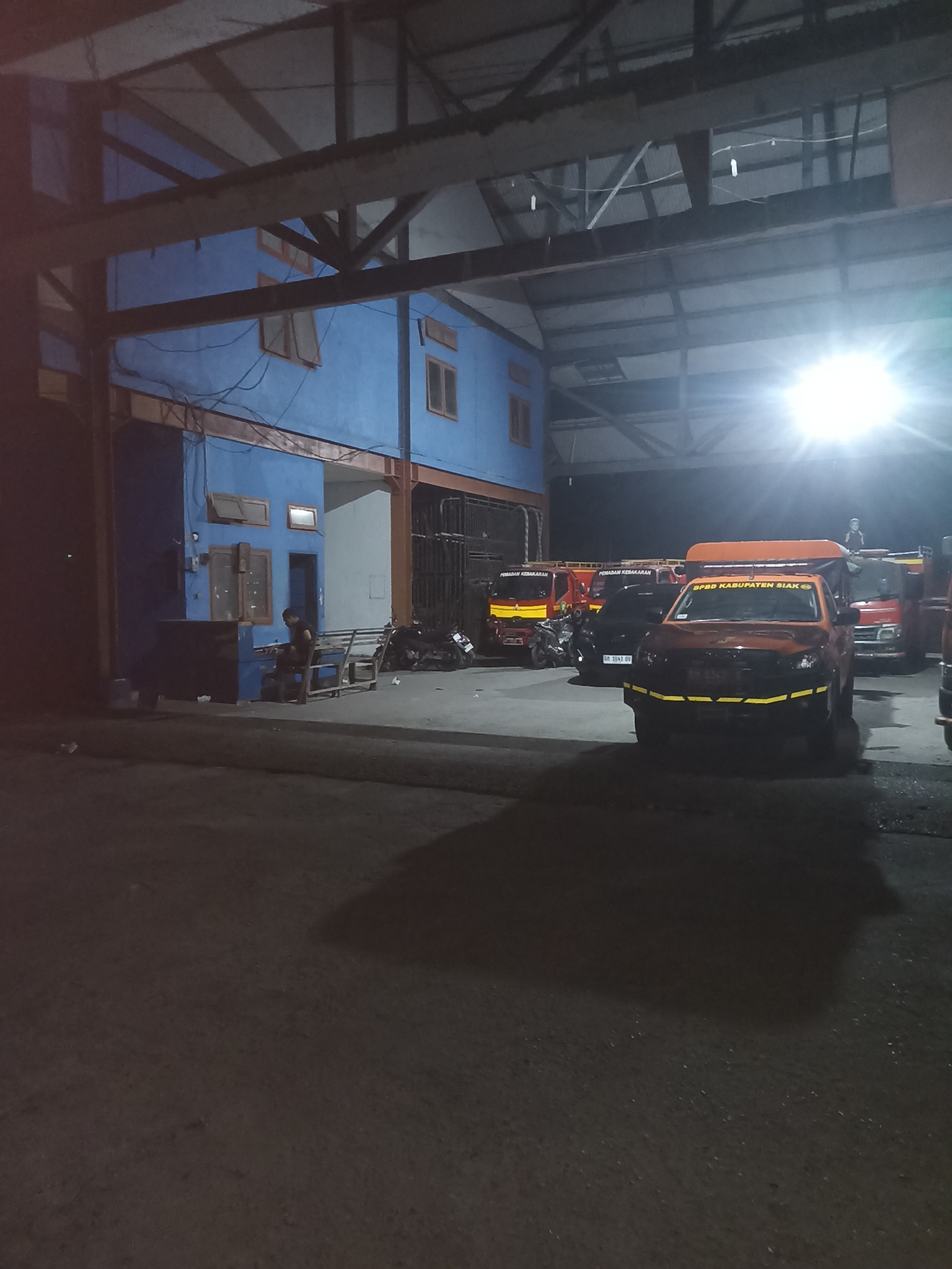 The image depicts an outdoor scene at night, likely in Kampung Tengah, Indonesia, with a bright overhead light illuminating the area. A blue building with orange-trimmed windows and doors stands on the left side of the frame. In front of the building, a dark blue cabinet is placed on the ground, and a person is seated on a wooden bench, appearing to interact with the cabinet. Several vehicles are parked in the area. In the foreground, an orange pickup truck with yellow reflective tape on its bumper and side is prominently featured. Behind it, a black car with a visible license plate is parked. Further back, several red trucks with "PEMADAM KEBAKARAN" (Fire Department) written on them are visible. Motorcycles are also parked near the red trucks. The ground is paved, and shadows are cast across it from the vehicles and the building.