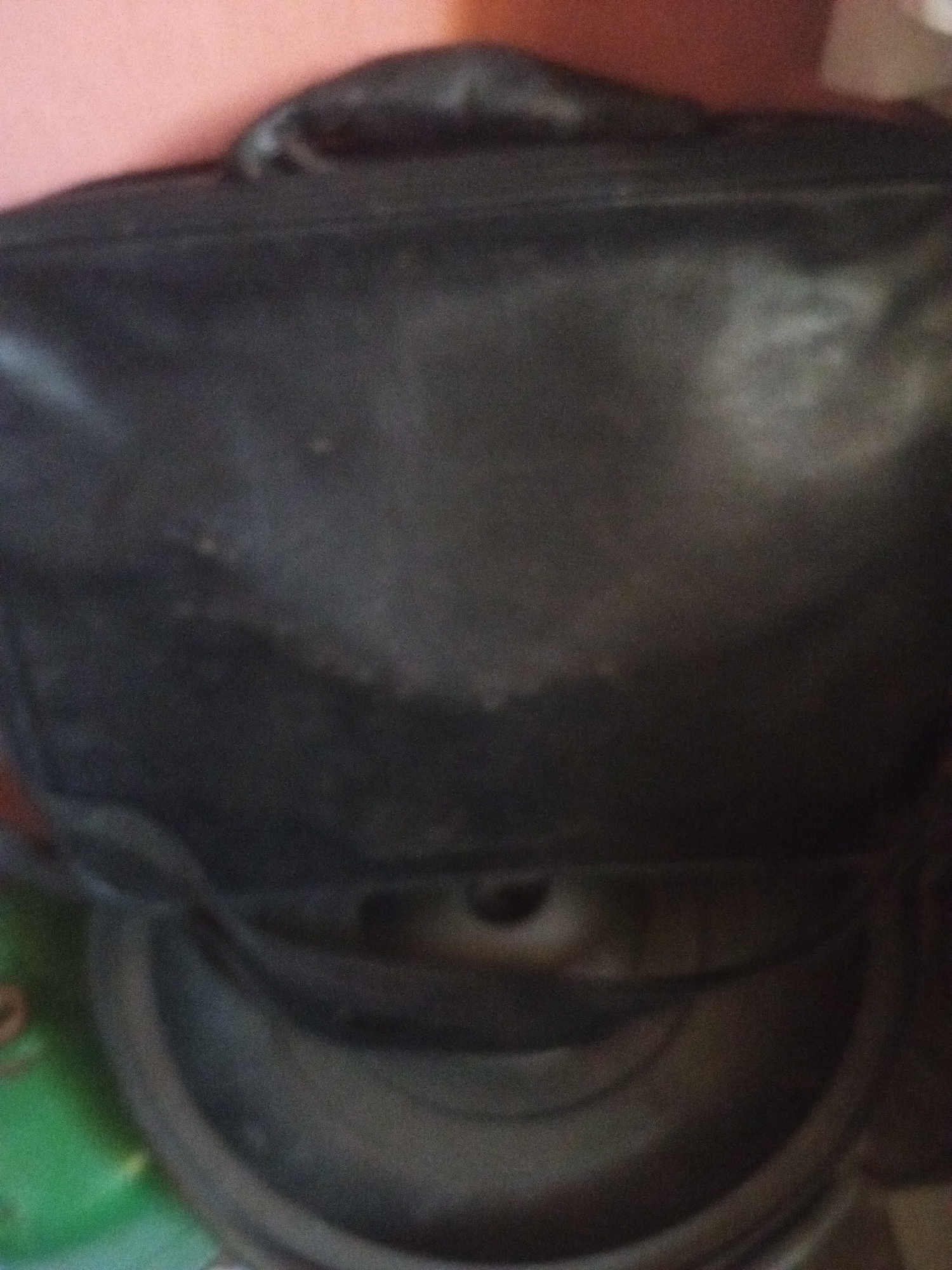 The image depicts a close-up, slightly blurry shot of a black bag, possibly a backpack or laptop bag, resting on top of a large speaker. The bag appears to be made of leather or faux leather and shows some signs of wear, with lighter patches on its surface. The background consists of an out-of-focus pinkish surface. The speaker itself is dark and has a textured surface, with visible concentric rings. To the left of the speaker, a portion of a green object is visible. The scene appears to be indoors, possibly in a domestic setting or a small business. There are no people or discernible landmarks that would indicate a specific location within Jalingo, Nigeria. The image lacks any specific details that would allow for a precise forensic description of the surroundings or the bag's contents.