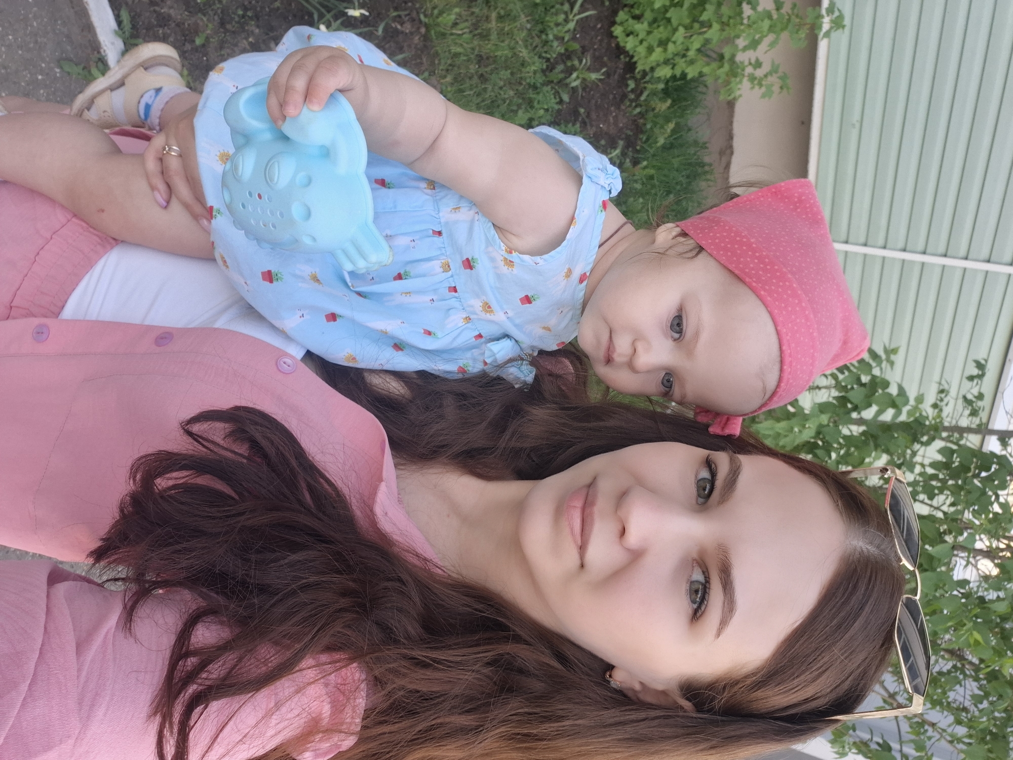A woman and a baby are outside, likely in a residential area in Chaikovsky, Russia. The woman has long brown hair, light skin, and is wearing a pink shirt. She is holding the baby, who has fair skin, a pink head covering, and is wearing a light blue dress with a strawberry pattern. The baby is holding a light blue plastic toy. A pair of sunglasses is visible near the woman's hair. The background shows green vegetation and a light green and white striped wall, possibly a fence or building.