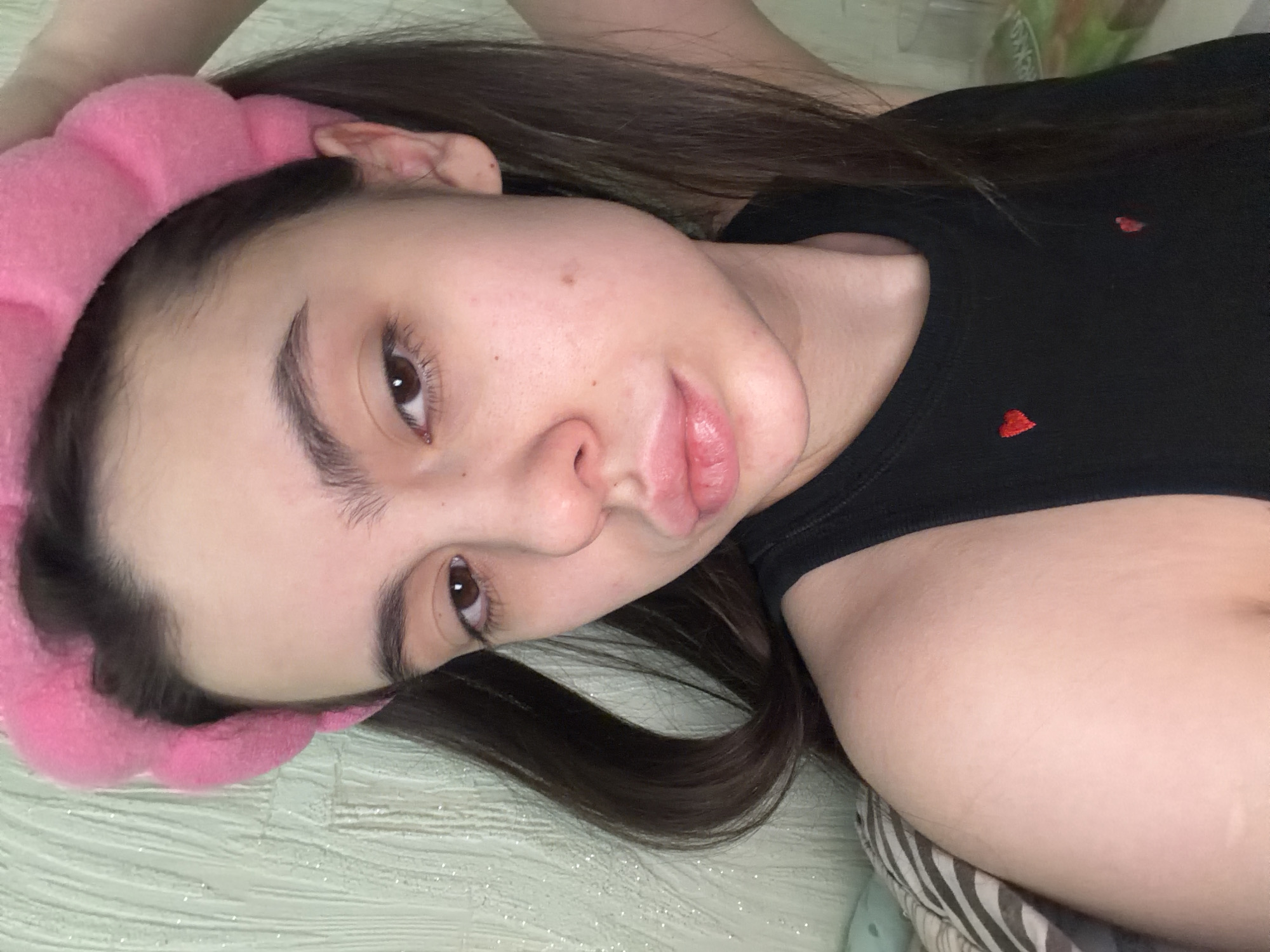 A young woman with brown hair and a pink headband is lying down. She is wearing a black top with small red heart details. The background is a light green surface with some sheen. The photograph was taken in a private setting. There is no indication of it being located in the city of Chaikovsky, Russia.