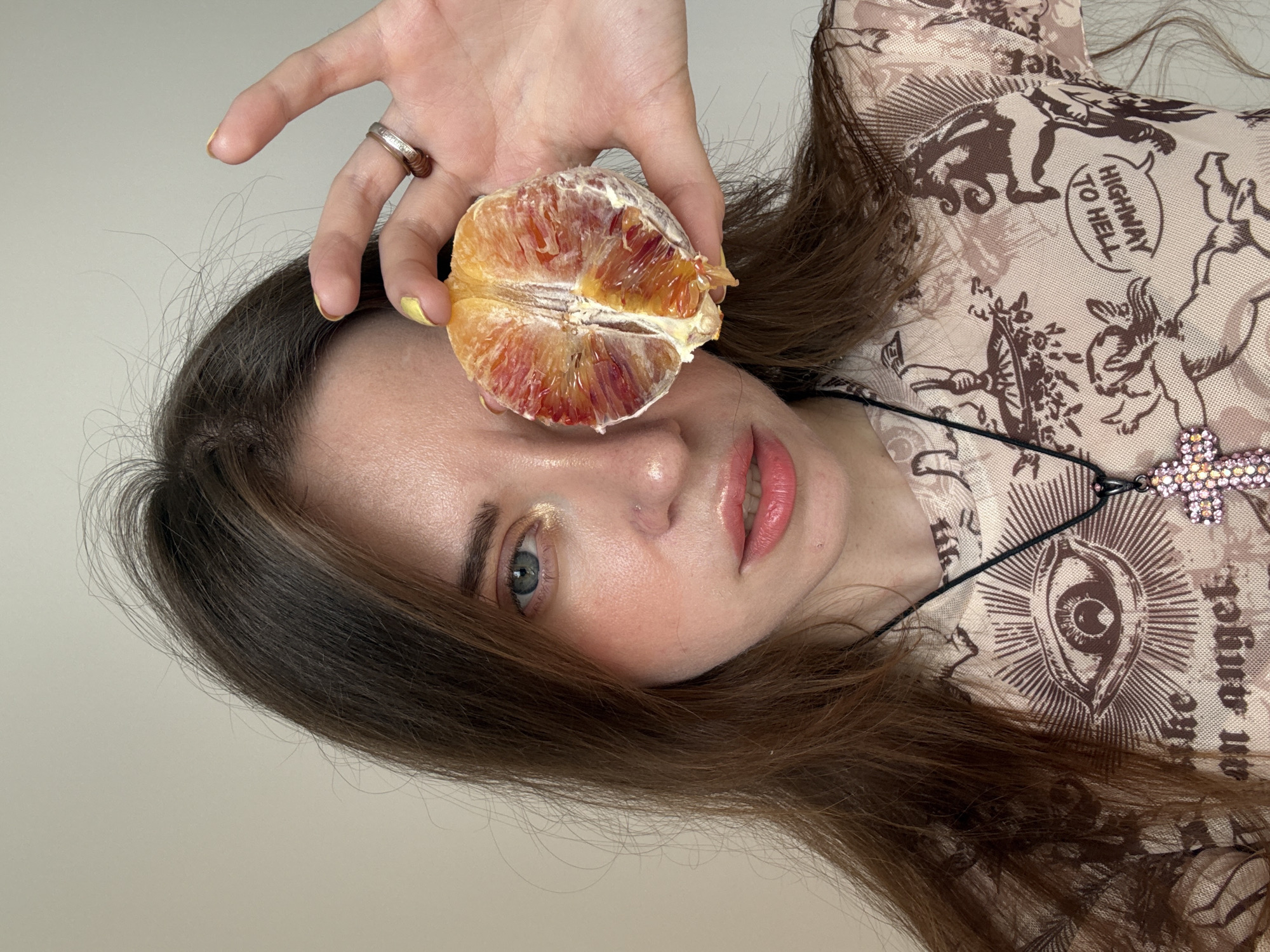 A young woman poses for a selfie indoors. She is holding a cut blood orange in front of her face, her hand displaying a ring and yellow nail polish. She has fair skin, light eyes, and brown hair. She wears a patterned blouse and a necklace with a cross pendant. The location appears to be indoors, with a plain wall in the background. There is no indication of Moscow, Russia.