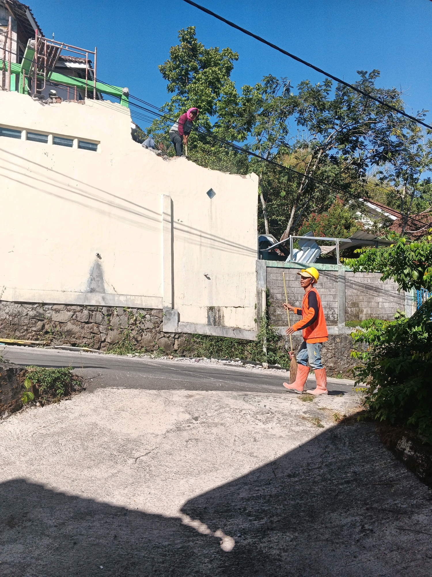 An outdoor medium shot captures two construction workers on a bright, sunny day in Keji, Indonesia. On the left, a worker in a pink headscarf and a dark shirt is on top of a partially demolished cream-colored building, using a shovel. Another construction worker, wearing an orange vest over a dark shirt, blue jeans, and bright orange boots, stands in the foreground on the right, holding a broom. He is looking towards the left and appears to be walking. The building appears to be undergoing renovation or demolition, with exposed beams and debris visible on the roof. The background consists of lush green trees and a clear blue sky. A paved road slopes downwards in front of the building, and a concrete area is visible in the lower foreground. The scene suggests ongoing construction or demolition work in a residential area.